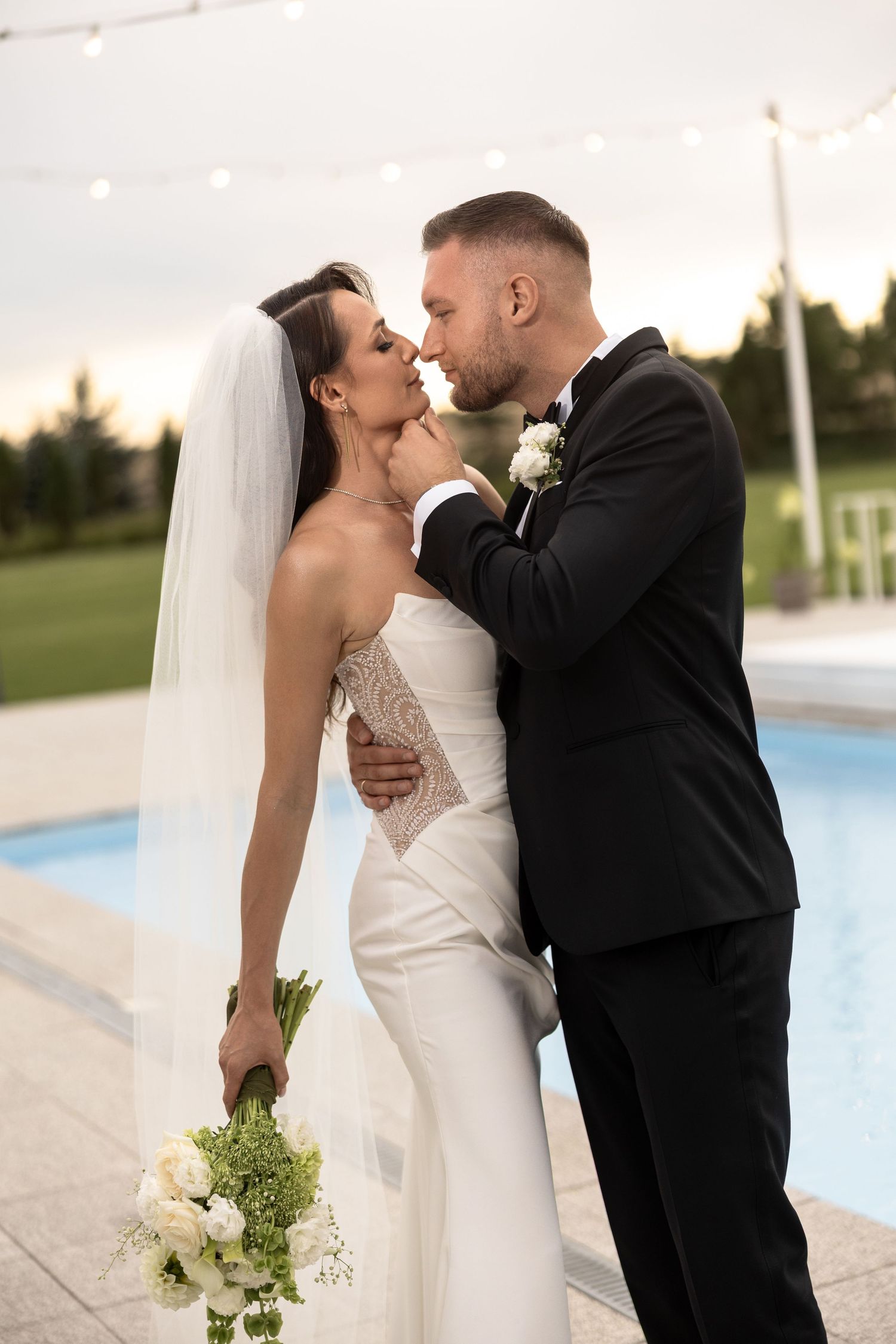 A bride and groom share a romantic kiss by a pool at dusk, she in a white gown with veil, he in a black tuxedo.