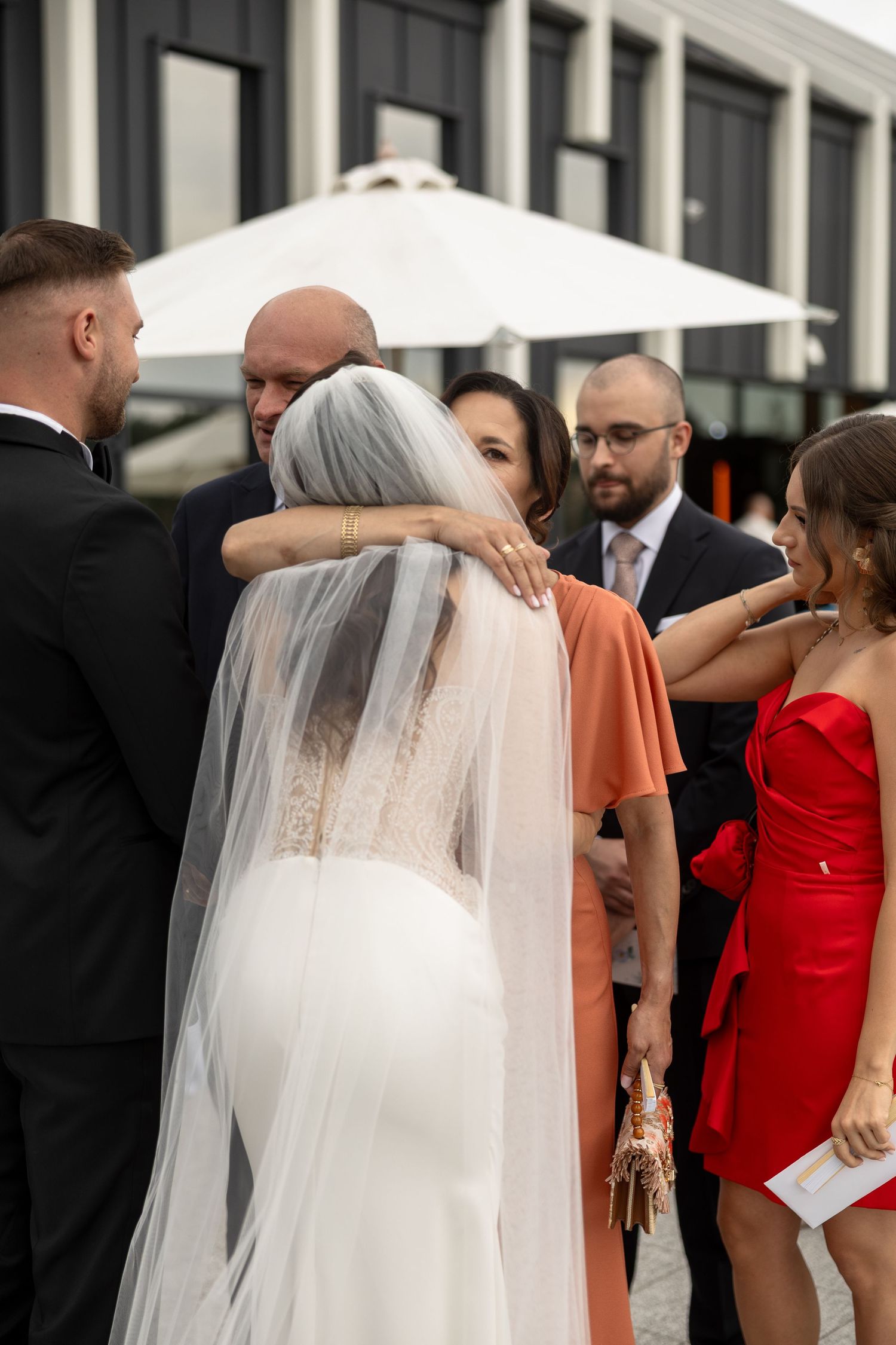 Bride in white gown and veil receiving hugs from wedding guests outdoors, with woman in red dress nearby.