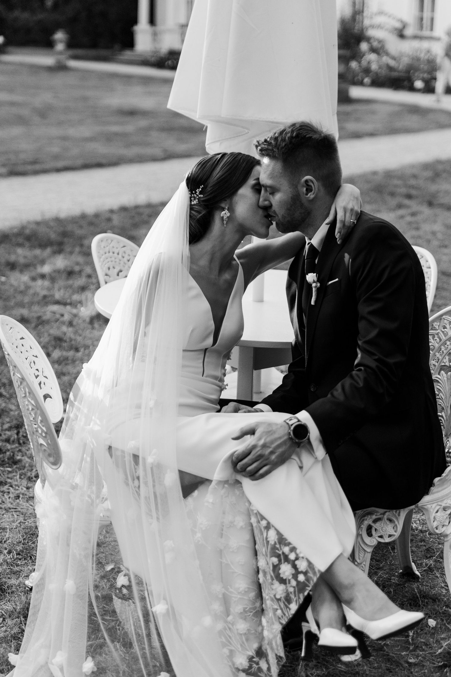 Black and white photo of a romantic wedding couple sharing a kiss while seated outdoors on vintage chairs.