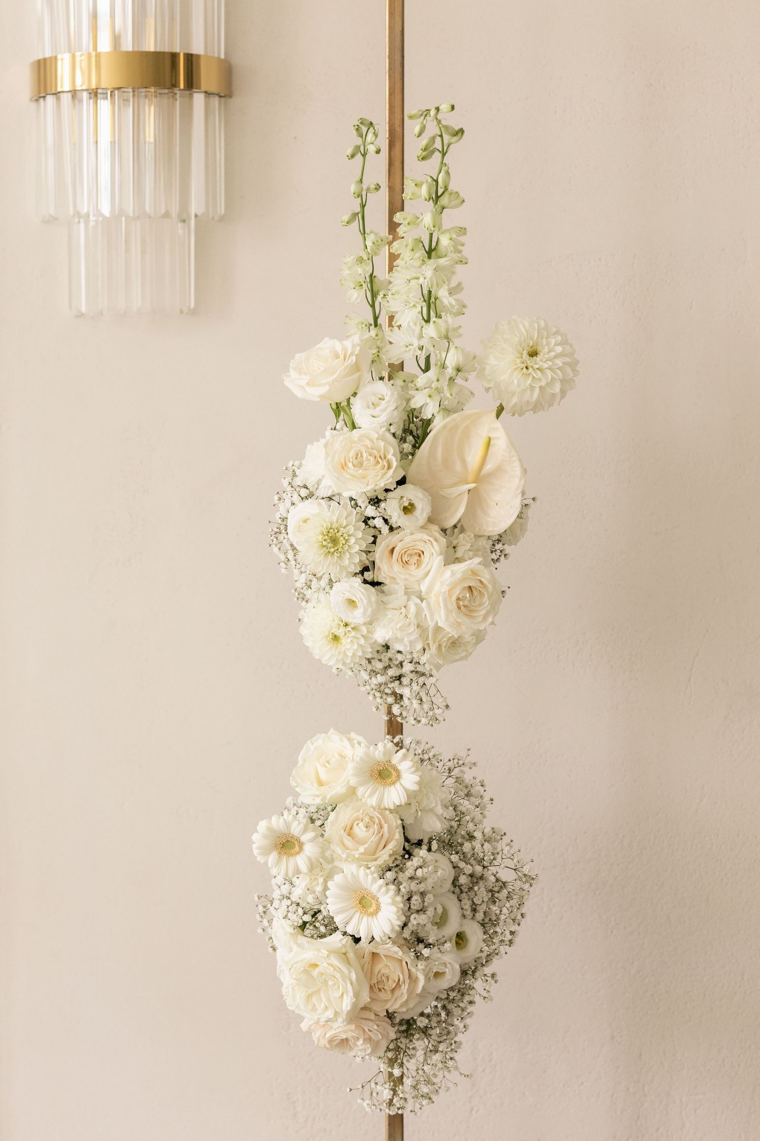 Elegant white floral arrangements with roses and baby's breath cascade down a light beige wall with modern gold sconces.