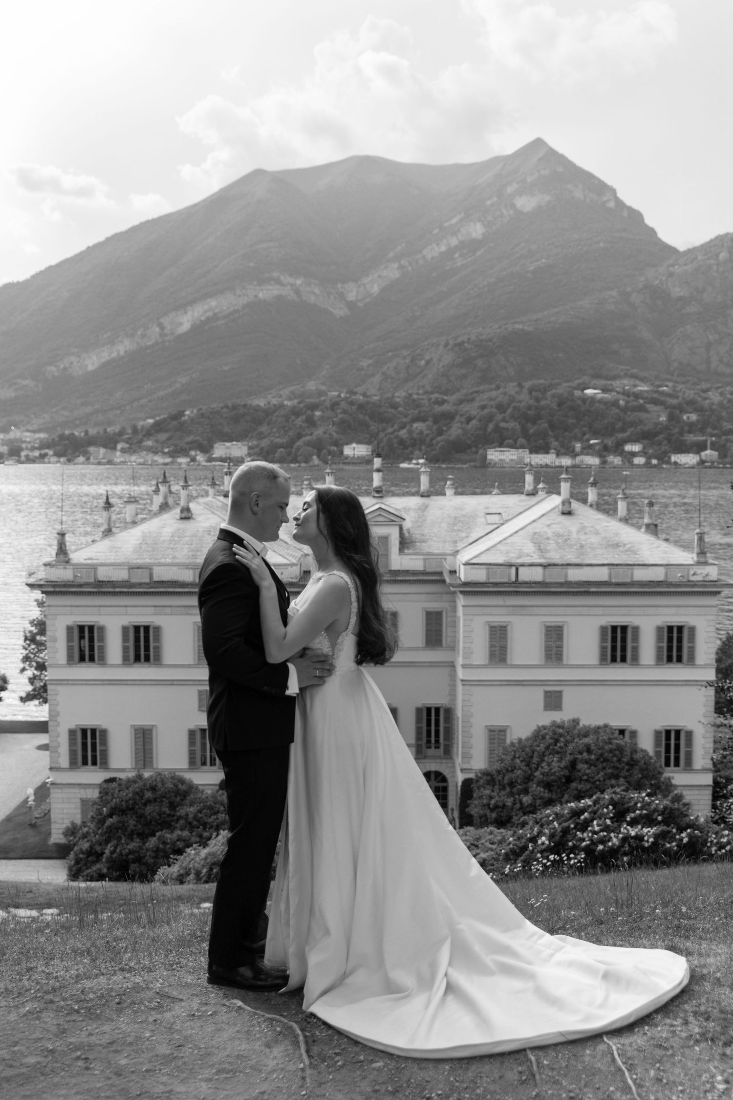 Black and white wedding photo of couple embracing before a grand villa with mountain and lake backdrop.