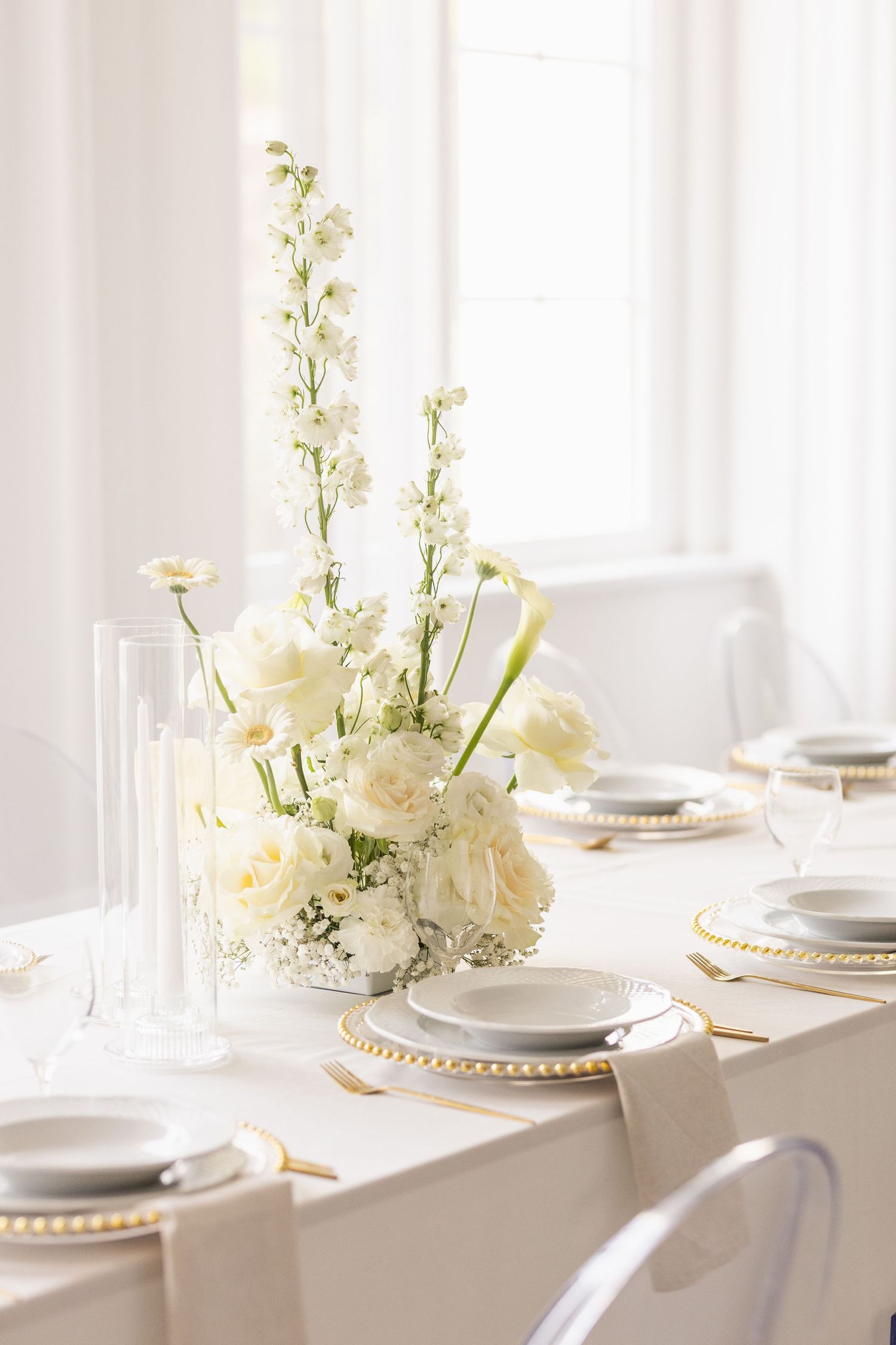 Elegant white table setting with floral centerpieces and beaded charger plates in soft natural light.
