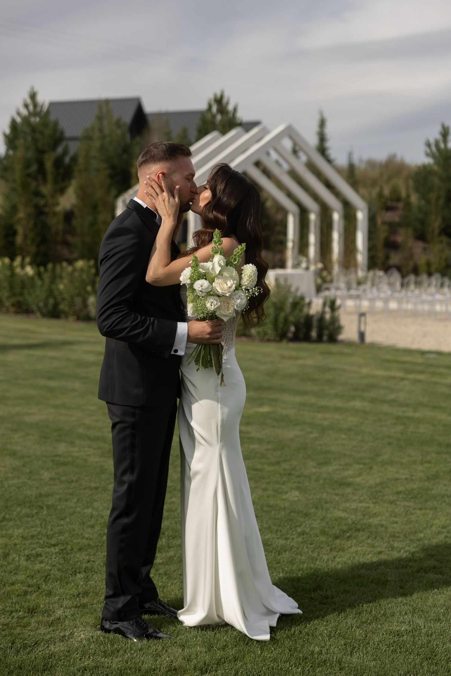 Bride and groom share a romantic kiss on green lawn, bride holding white rose bouquet, outdoor wedding venue backdrop.