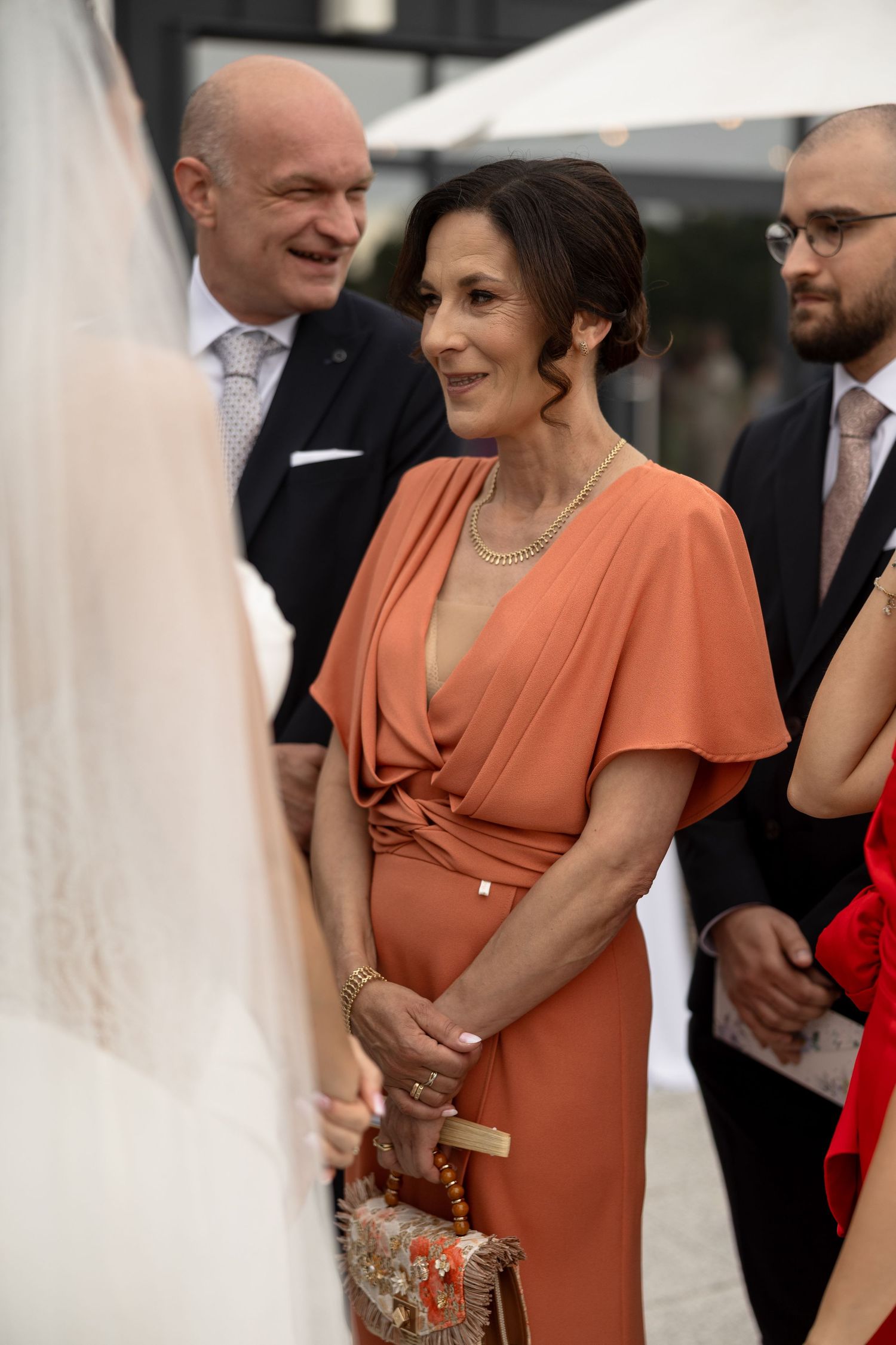 Woman in elegant orange dress smiling at a wedding ceremony, with guests in formal attire in the background.