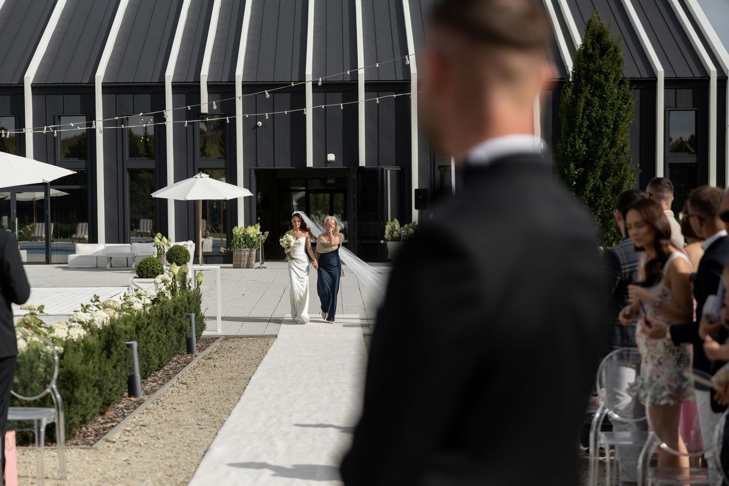 A bride walks toward her groom at an elegant outdoor wedding venue with modern architecture and lush greenery.