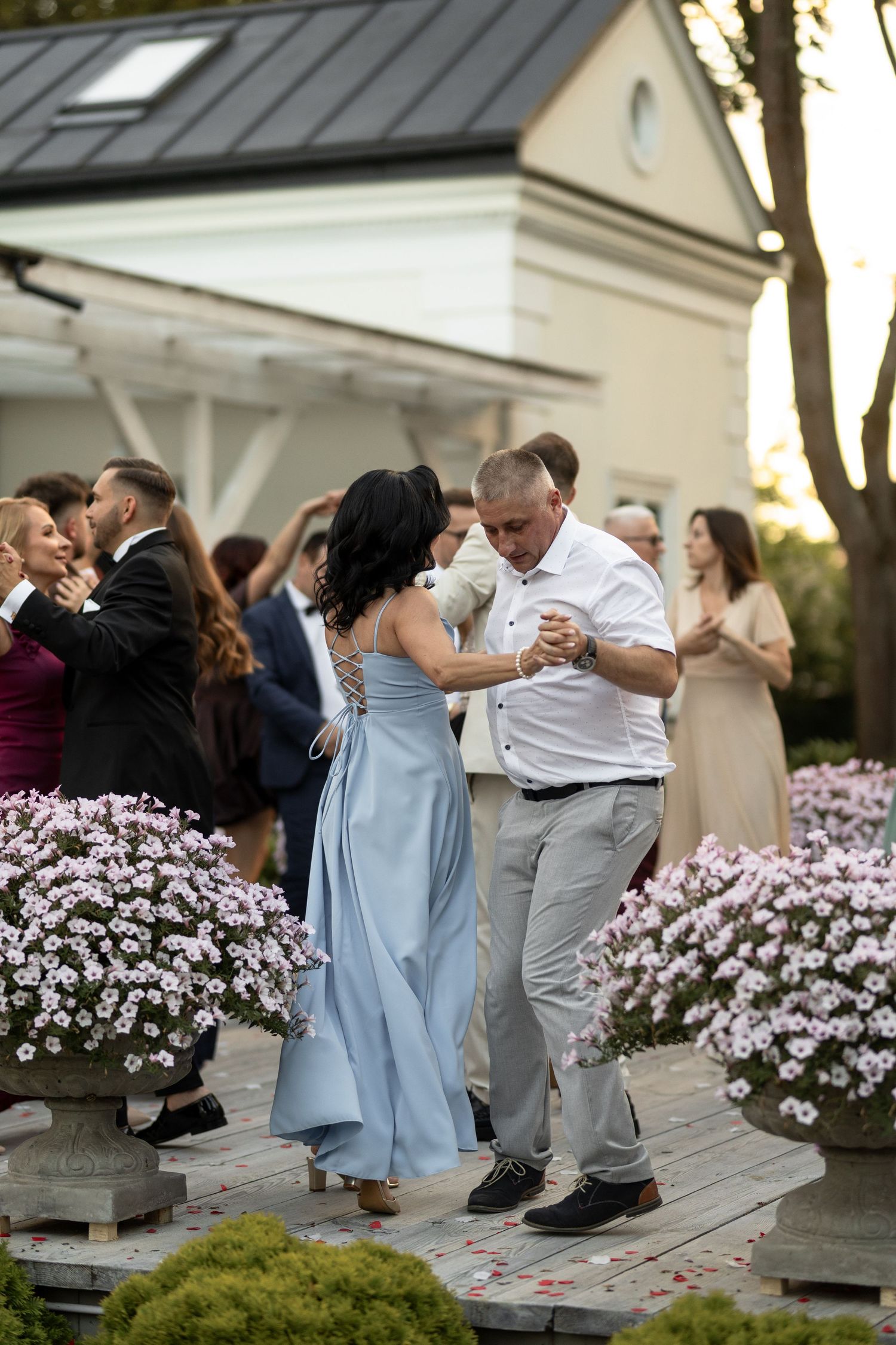 Couple dancing outdoors at a wedding reception surrounded by blooming flowers and guests near a white building.