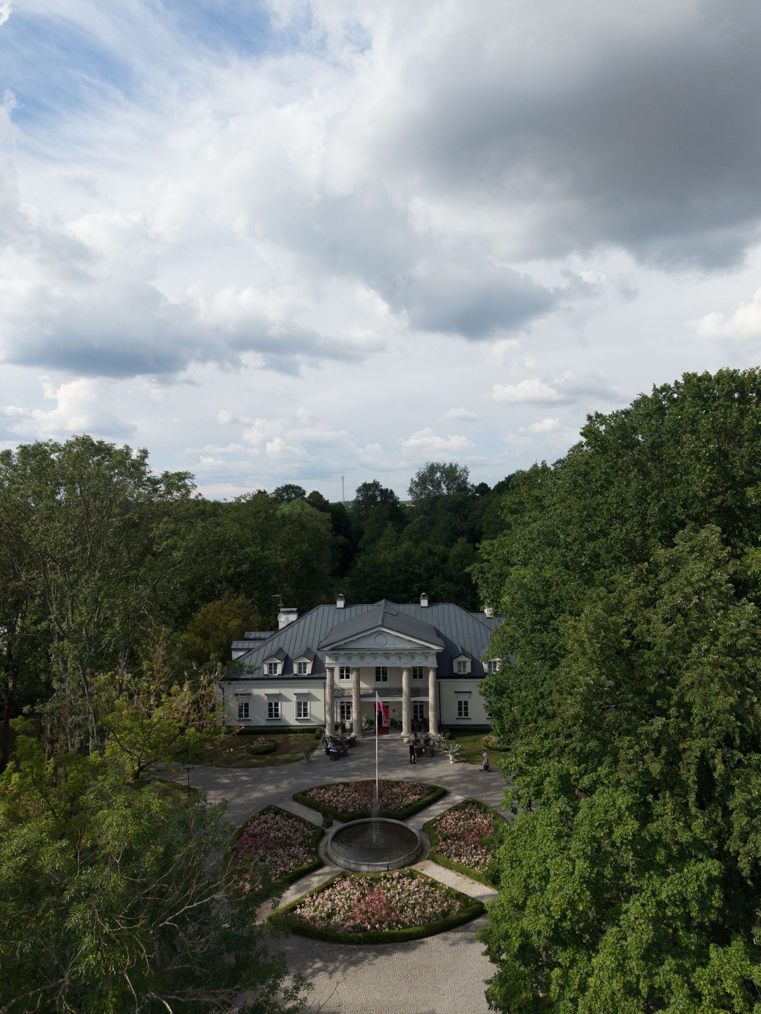 Aerial view of a grand white neoclassical mansion surrounded by lush green trees and a formal garden under a cloudy sky.