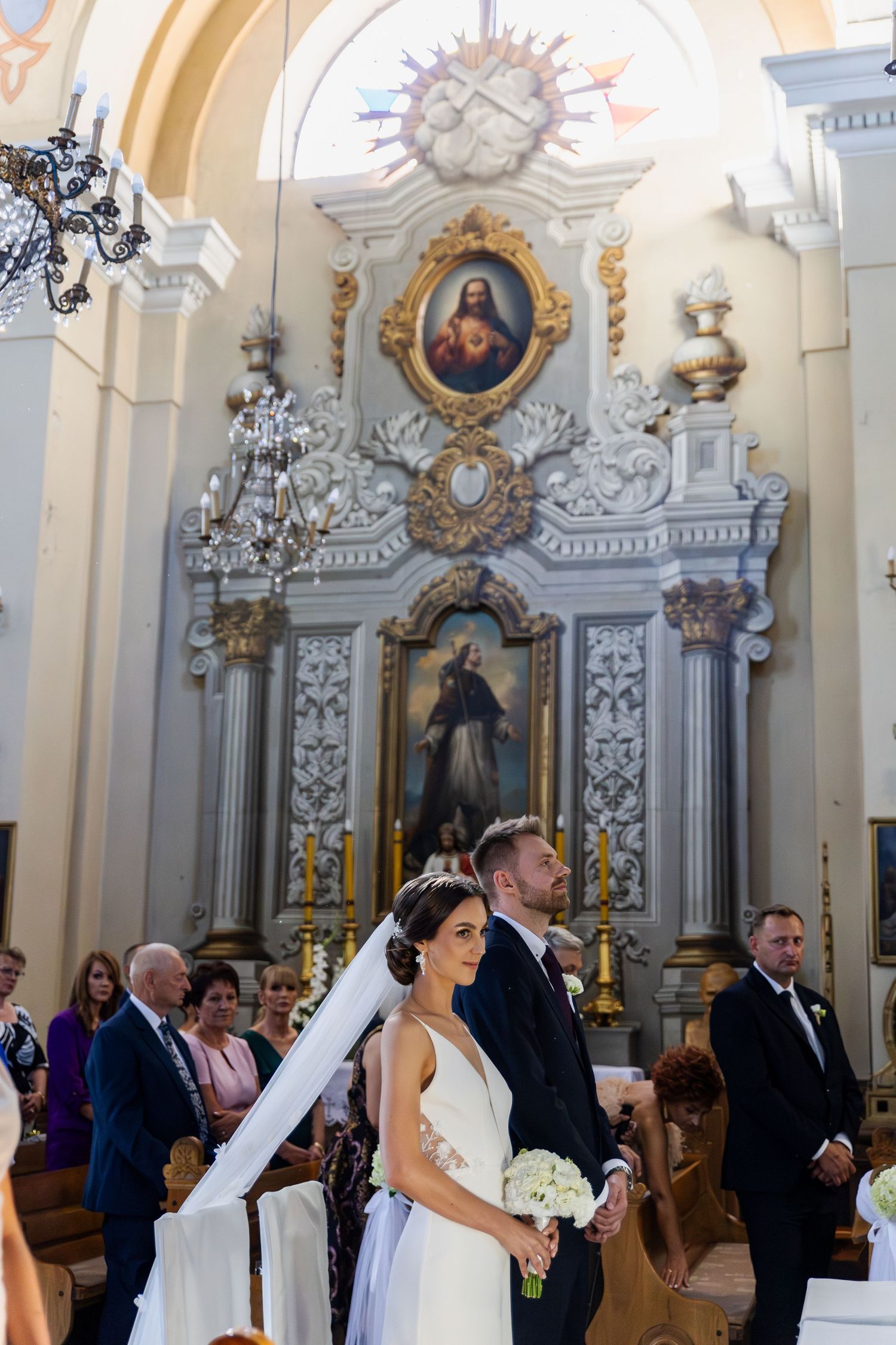 A couple stands at the altar in an ornate baroque church interior with gilded decorations and religious paintings.