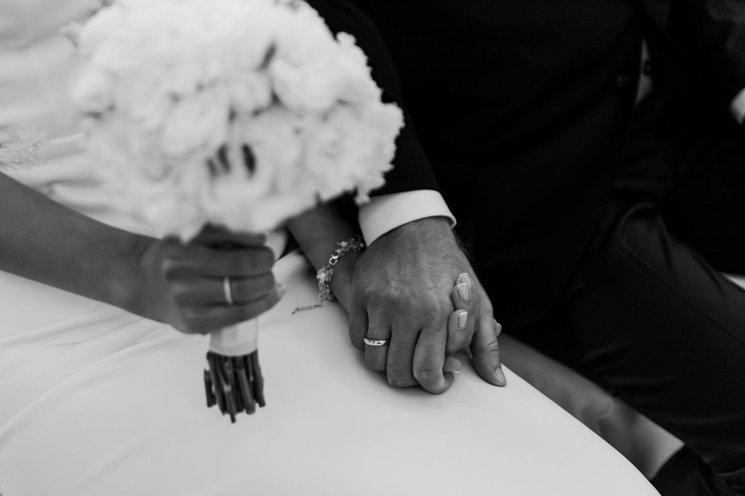 A black and white close-up of hands resting together on a white dress with jewelry details.
