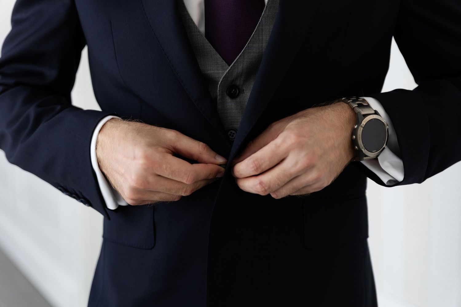 Close-up of hands buttoning a navy suit jacket over a dark sweater vest, with a wristwatch visible.