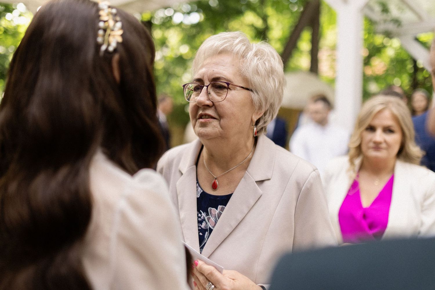 People gather outdoors for a social gathering with one wearing a beige blazer and another in bright pink clothing in background.