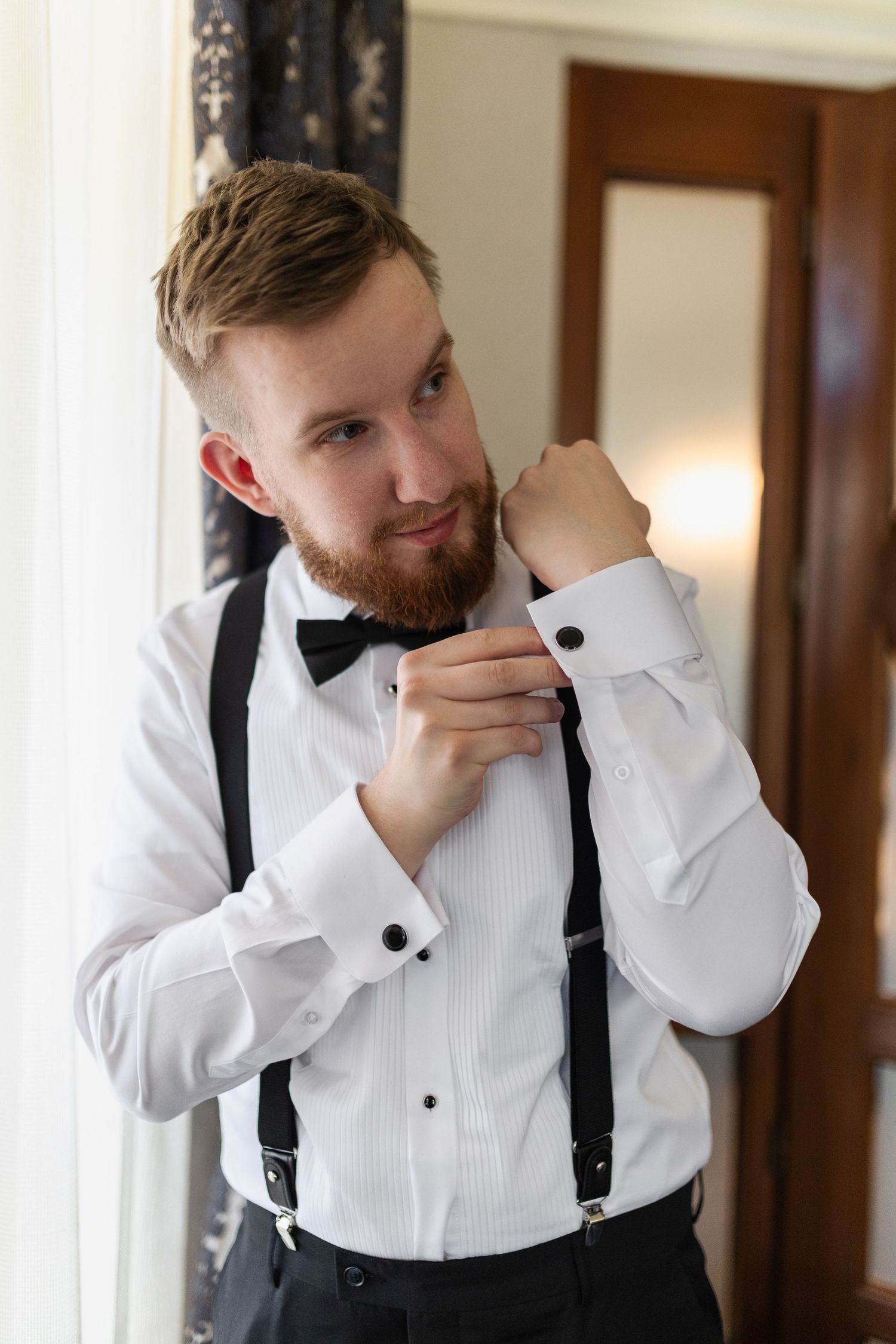 A groom adjusts his shirt cuff while wearing a classic white tuxedo shirt with black bow tie and suspenders.