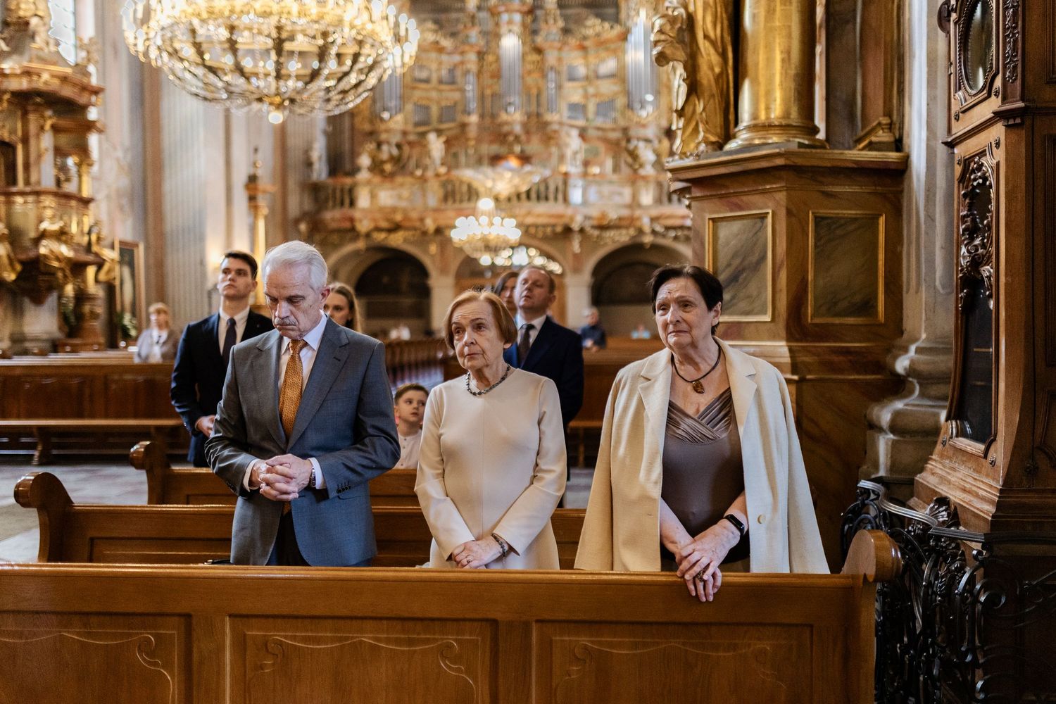 People gather in wooden pews at an ornate church with golden chandeliers and baroque architectural details.