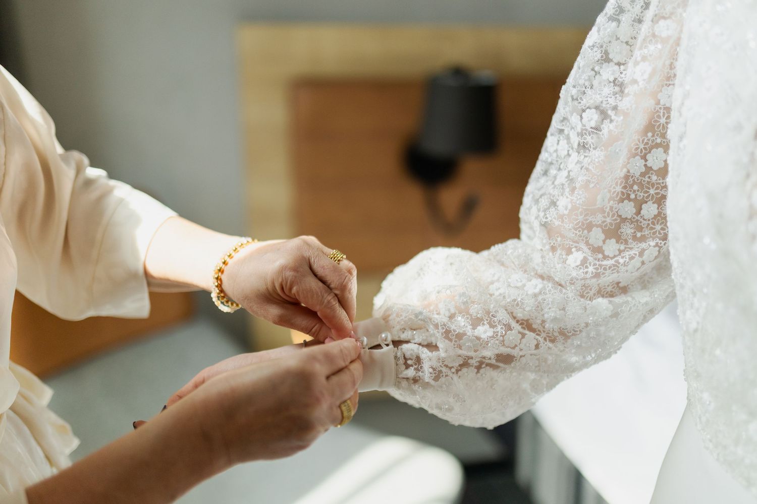 Hands carefully fastening delicate lace buttons on a white wedding dress sleeve.