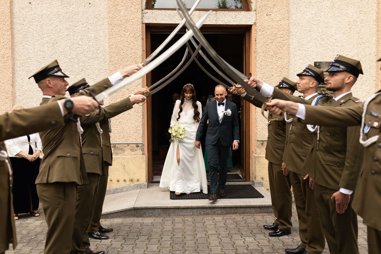 Uniformed military members hold up crossed swords in an archway as newlyweds exit a church building.