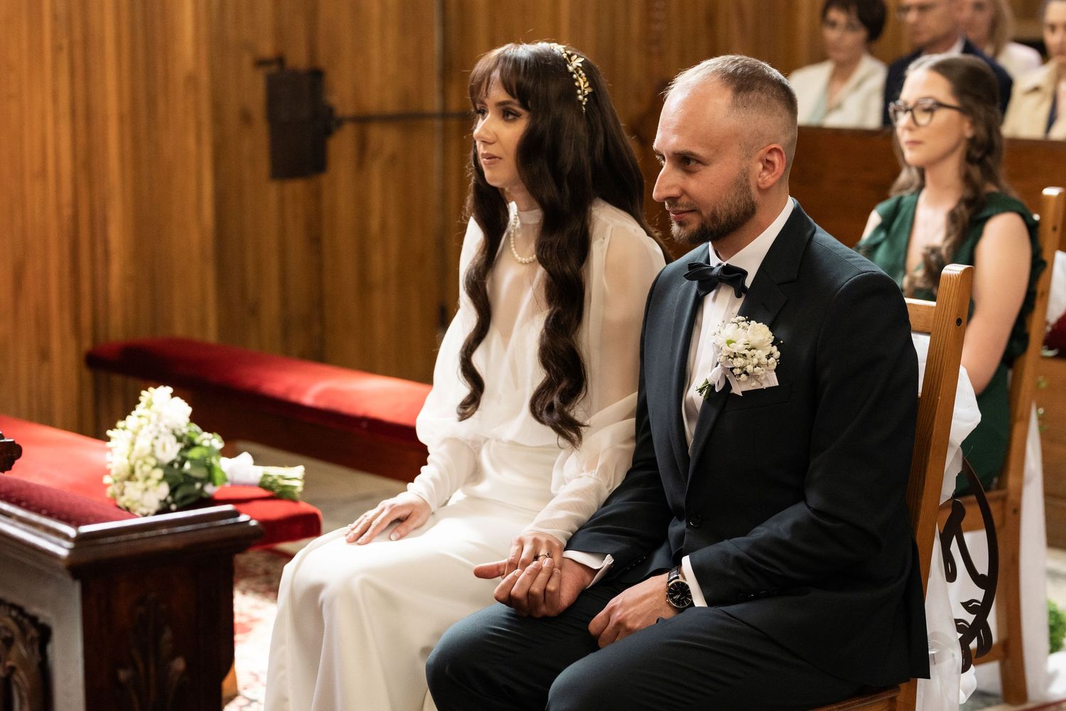 A bride and groom sit together during their wedding ceremony in a church with wooden paneled walls.