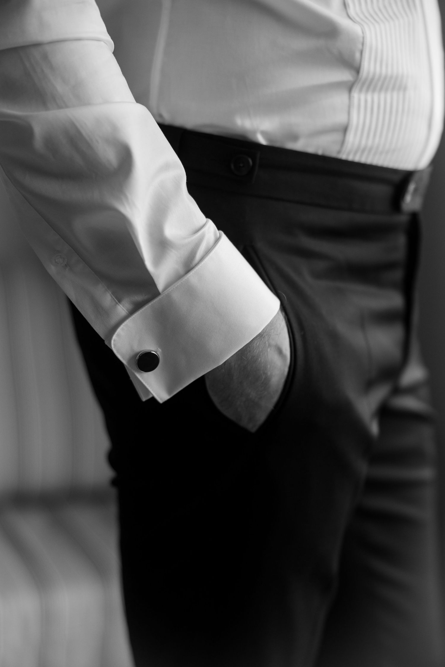 Black and white close-up of a man in a formal white dress shirt with cufflinks, hand in pocket of dark trousers.