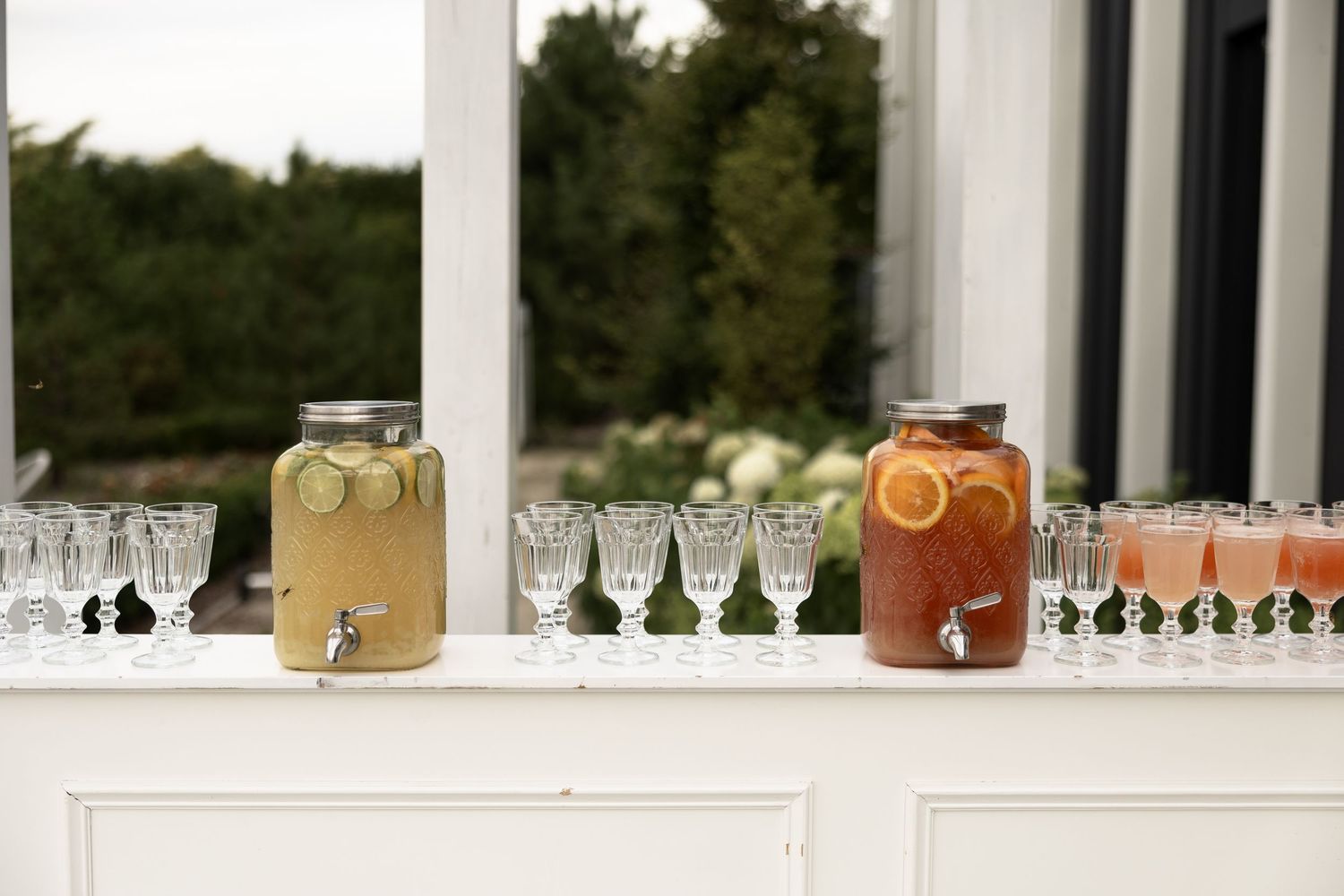 Two beverage dispensers with citrus fruit infusions flanked by crystal glasses on a white buffet table.