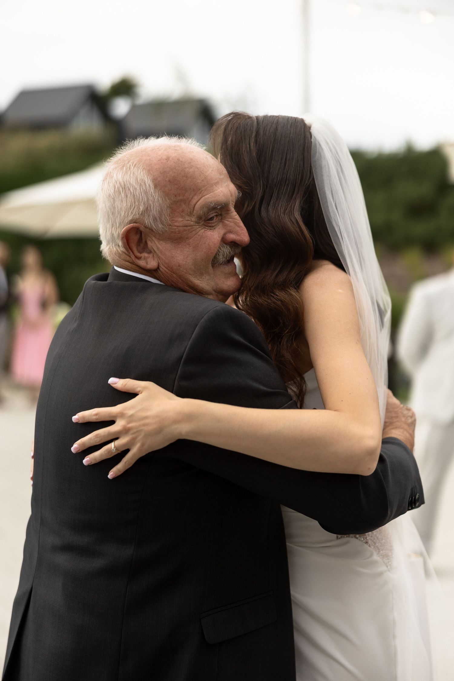 A bride in a white veil shares a heartfelt hug with an elderly man in a dark suit at a wedding.