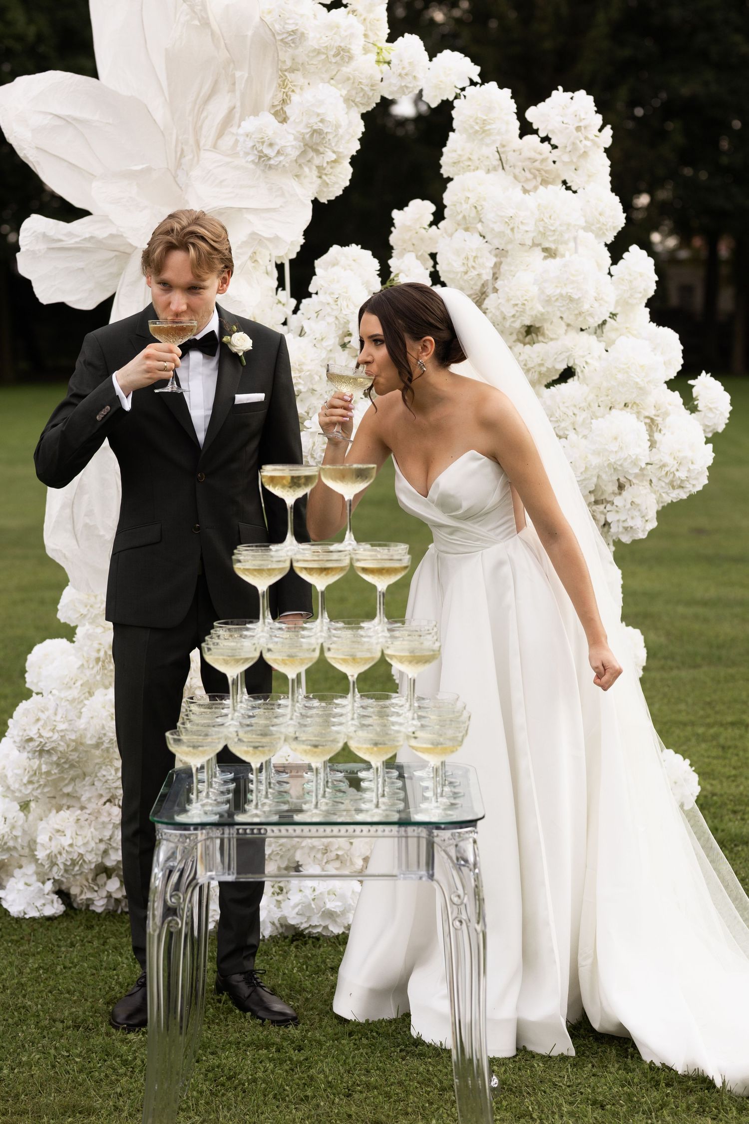 Bride and groom toasting champagne beside a pyramid of glasses, surrounded by white floral arch decor outdoors.