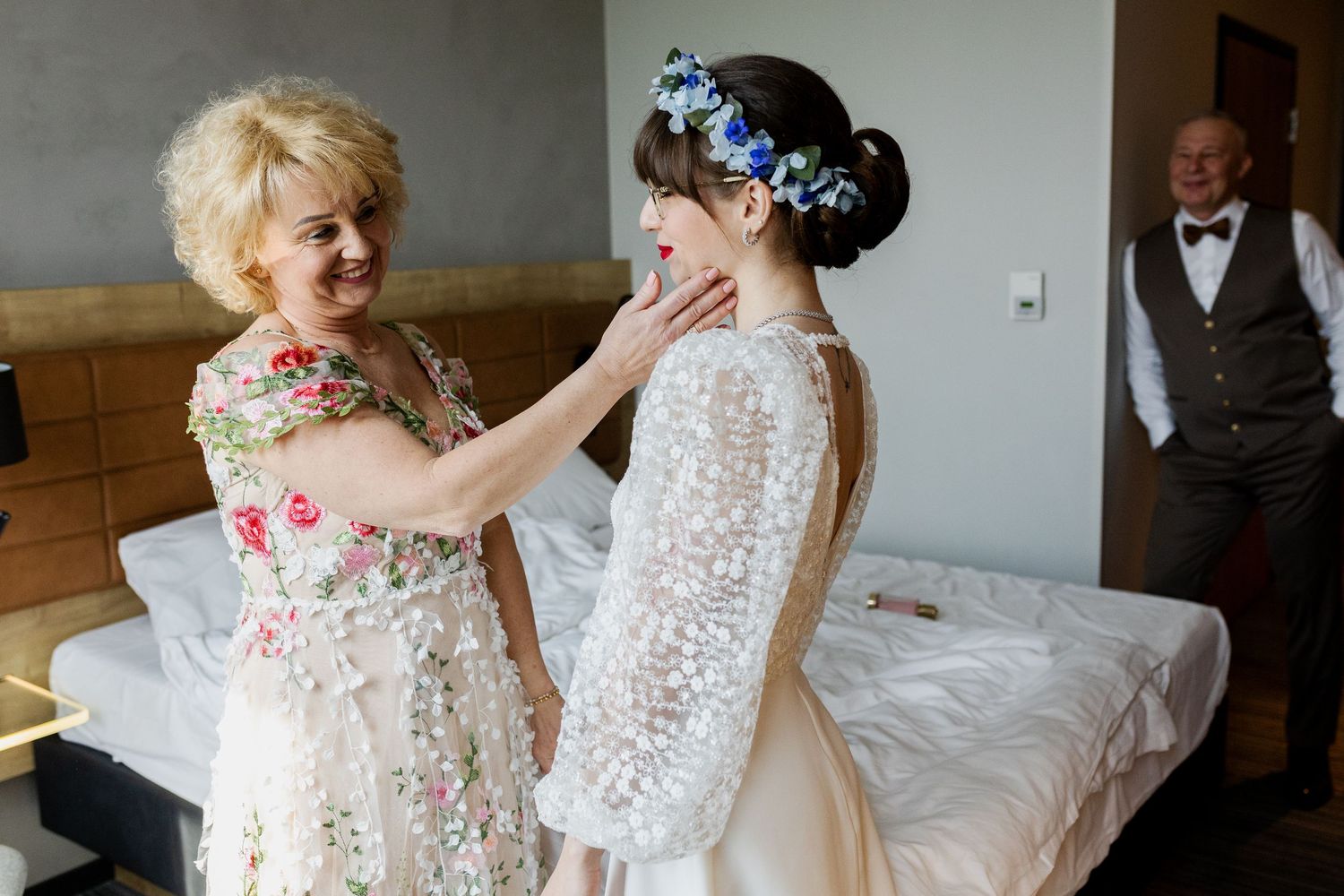 Two people share an emotional moment in a hotel room, one wearing a floral dress and the other in a white lace gown.