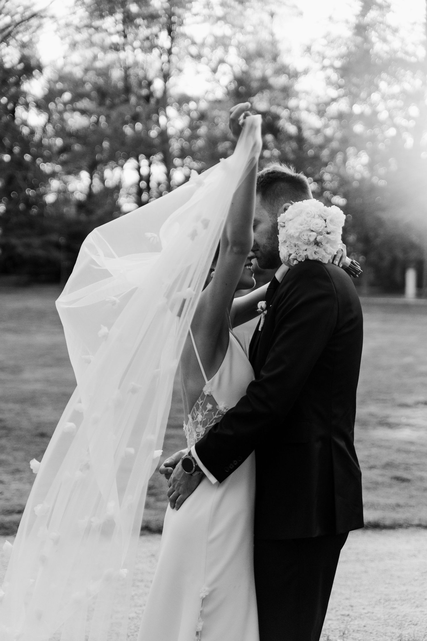 A romantic black and white wedding photo showing a flowing veil and tender kiss in a sunlit outdoor setting.