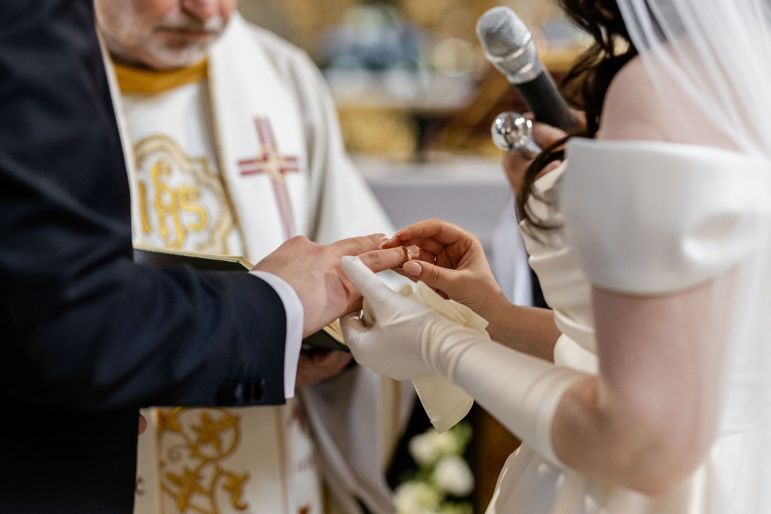 Two hands exchanging wedding rings during a ceremony with a priest in the background.