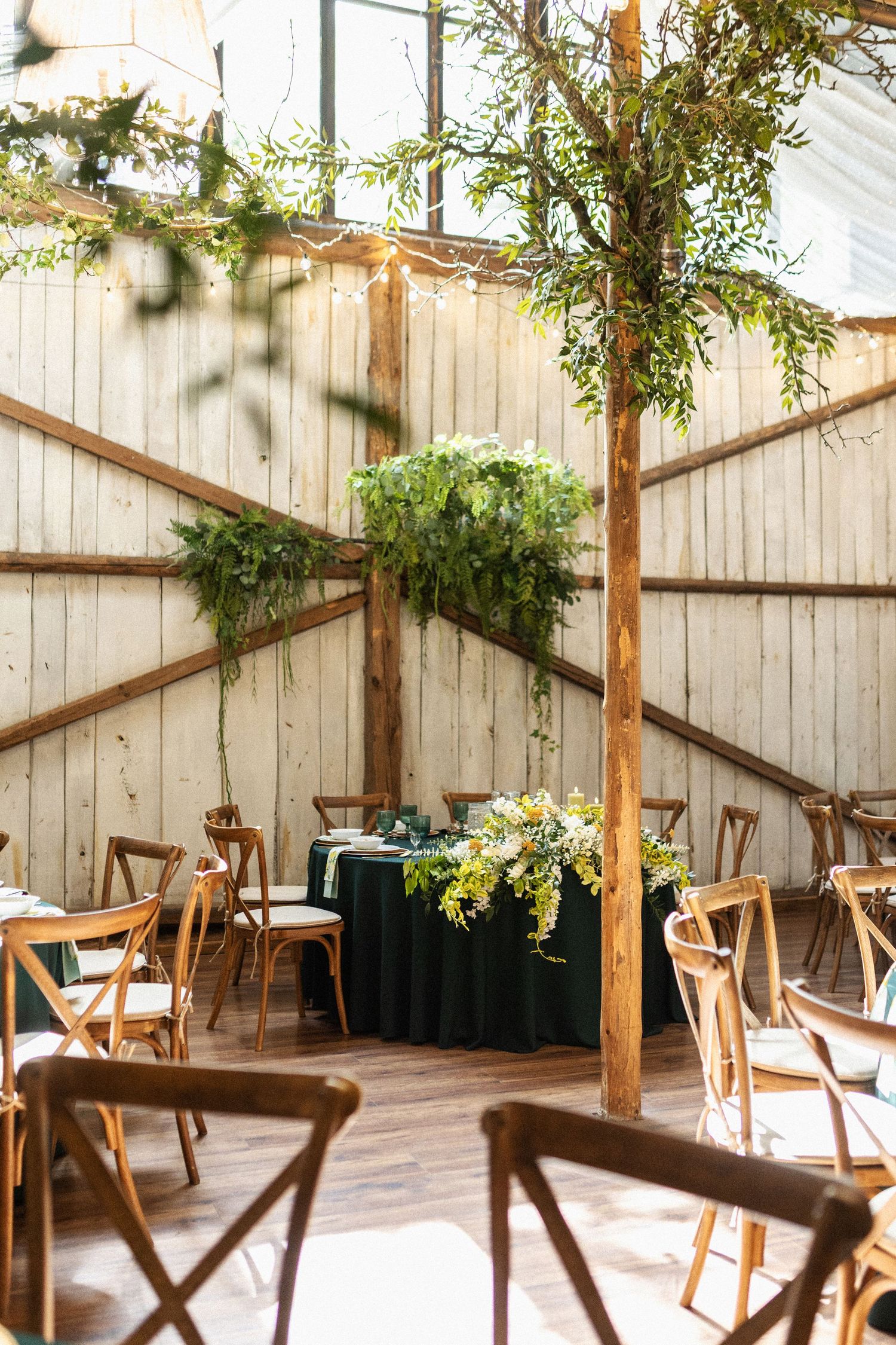 Rustic greenhouse wedding venue with wooden crossback chairs, greenery-adorned poles and dark tablecloth centerpiece.