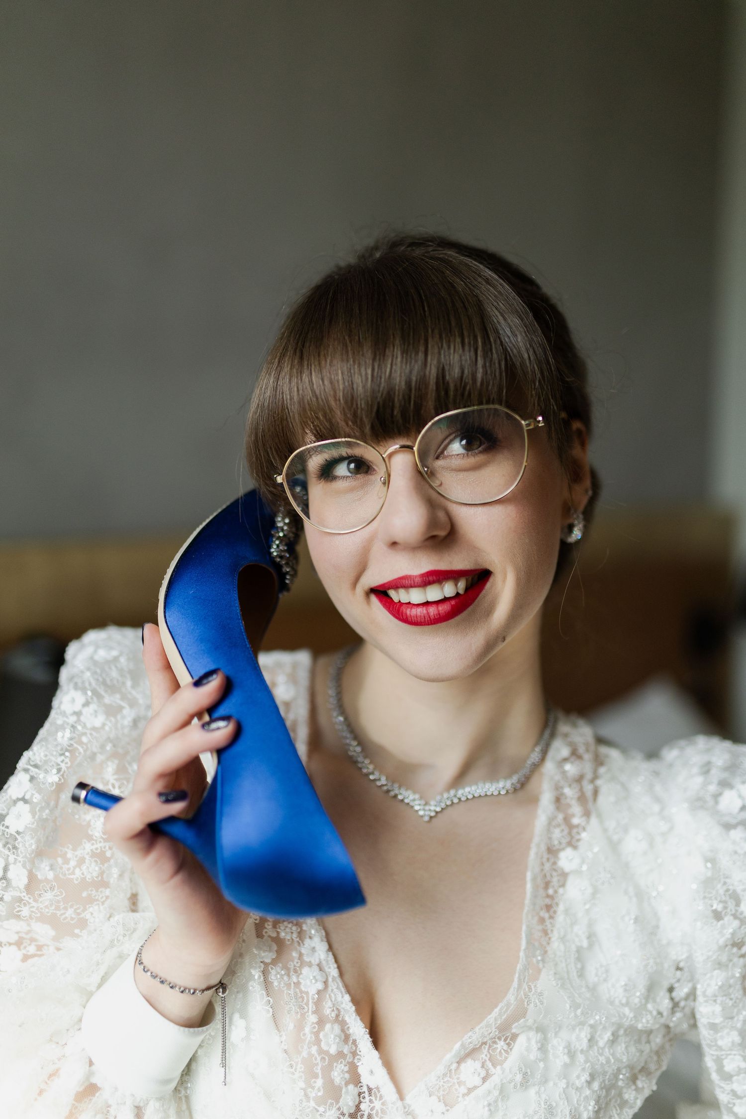 A bride in white lace and glasses holds a bright blue high heel shoe while wearing a sparkly necklace and red lipstick.