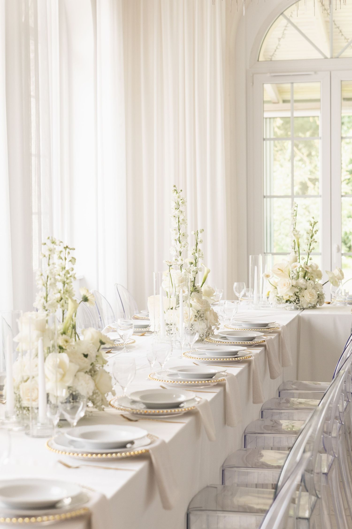 Elegant white wedding reception tables with crystal chairs and floral centerpieces in a bright sunlit room.