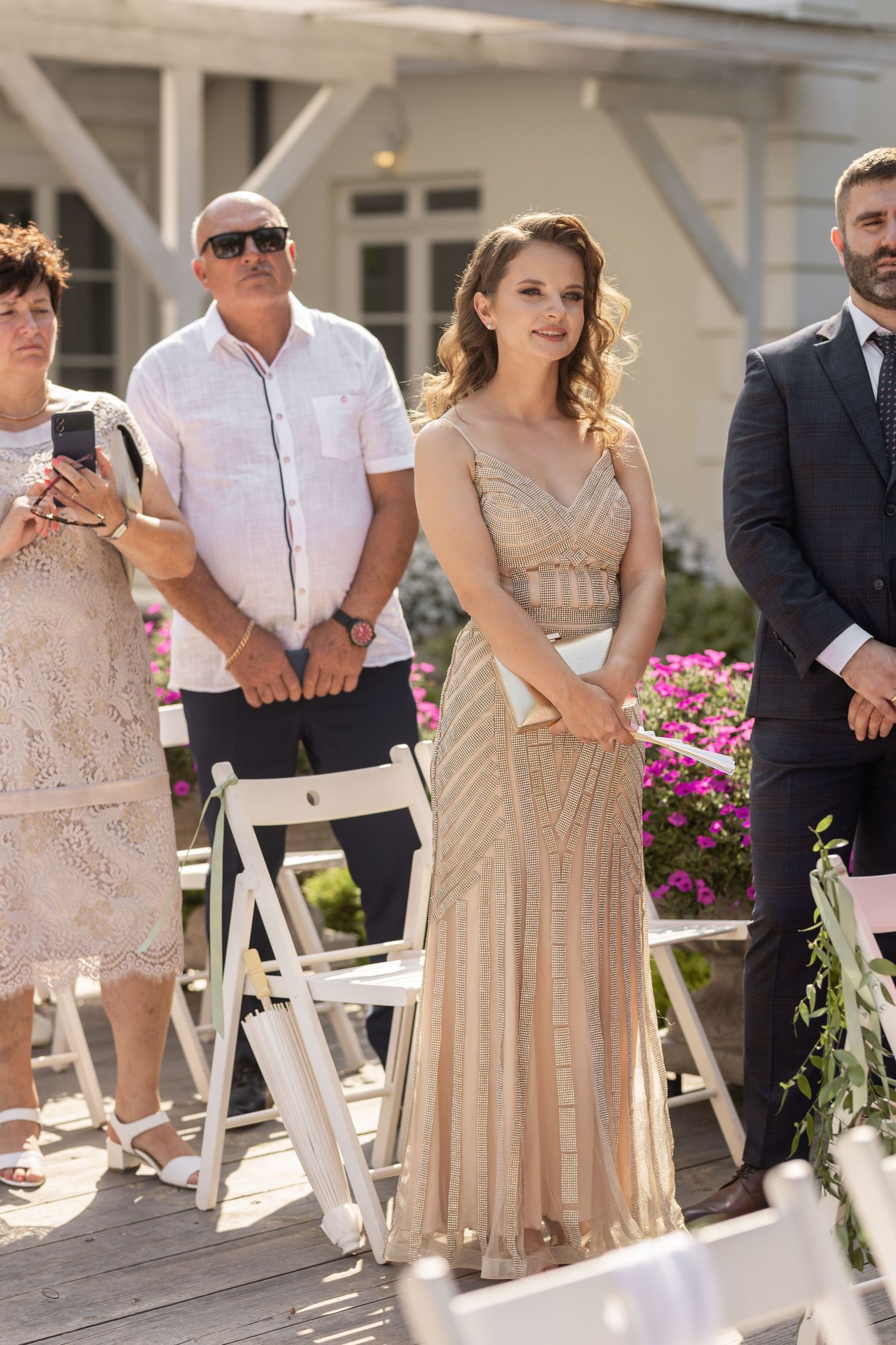 Wedding guests in champagne-colored dresses stand beside white folding chairs at an outdoor ceremony venue.