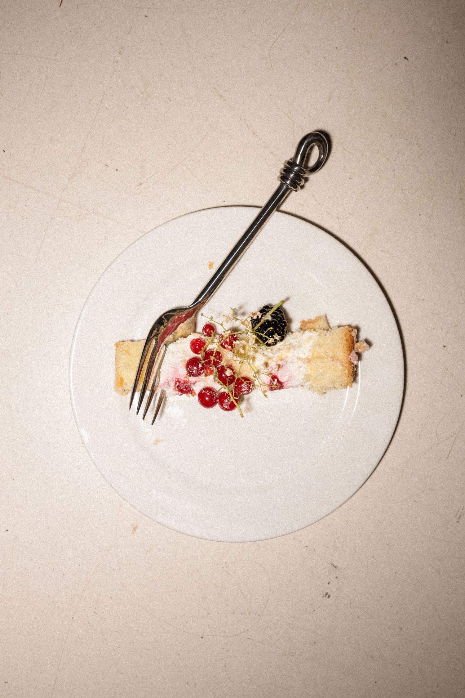 An ornate silver fork rests on a white plate with remnants of dessert and red currants.
