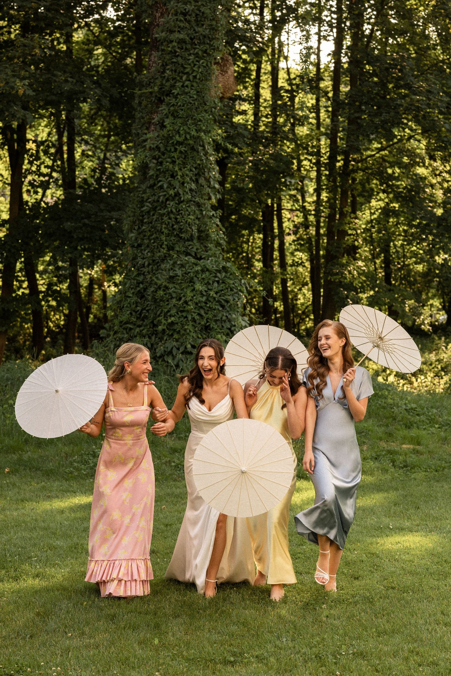 A group of friends in elegant dresses walk together on grass holding white parasols at a summer garden party.