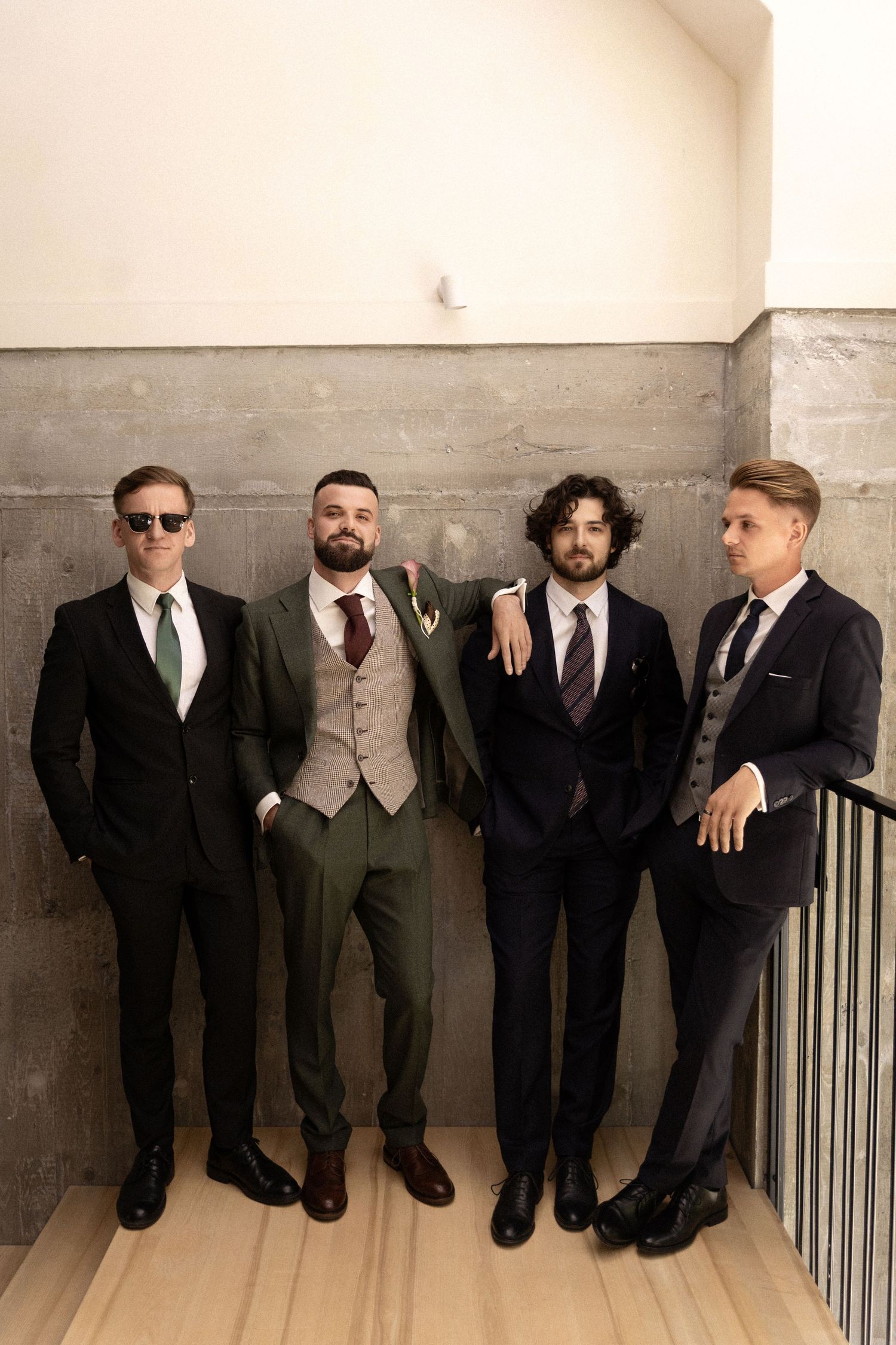 Four men in formal suits and ties standing against a wall, with one wearing a tan vest and another having curly hair.
