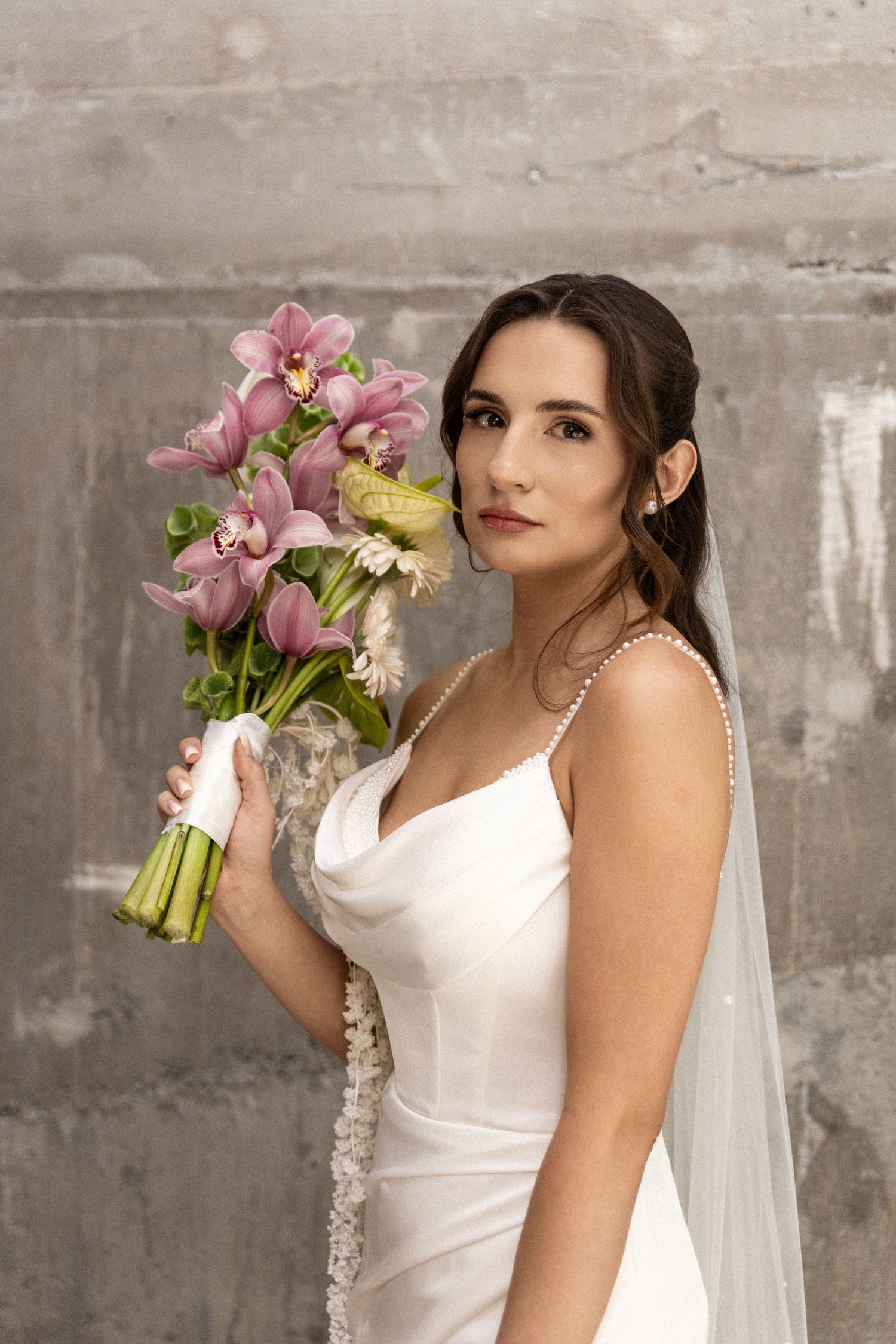 Beautiful bridal portrait with pink and yellow orchid bouquet against textured concrete wall.