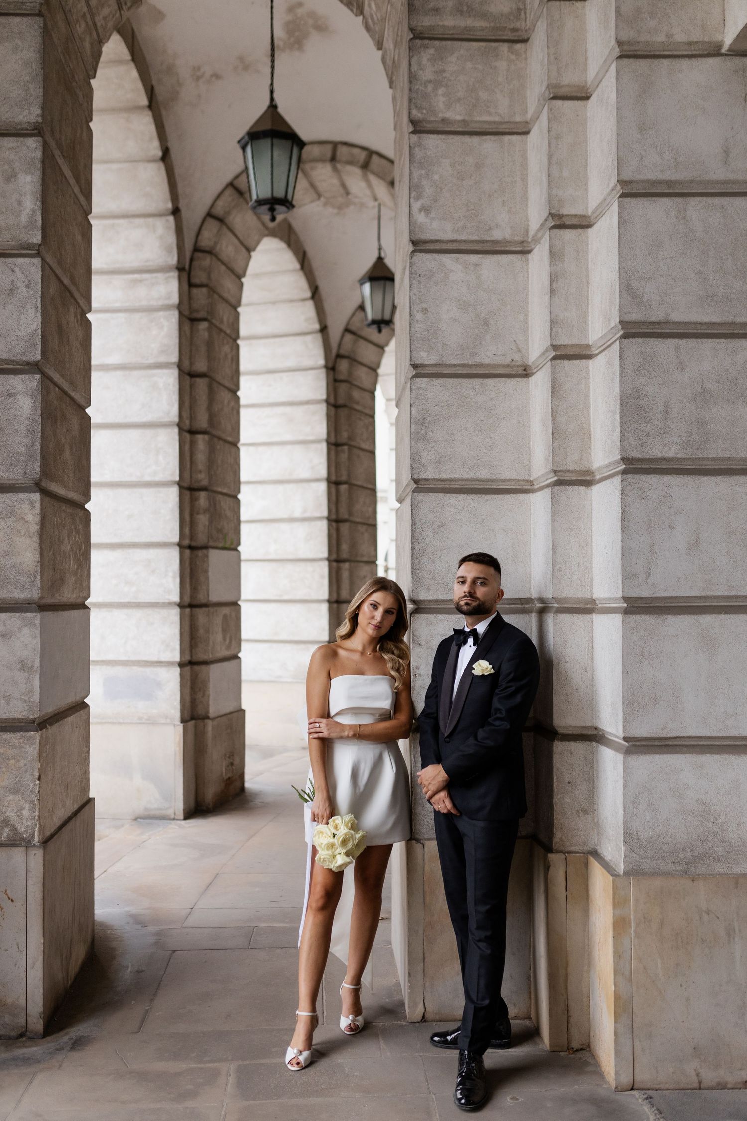 Elegant couple poses in formal wedding attire against a historic stone archway with lanterns.