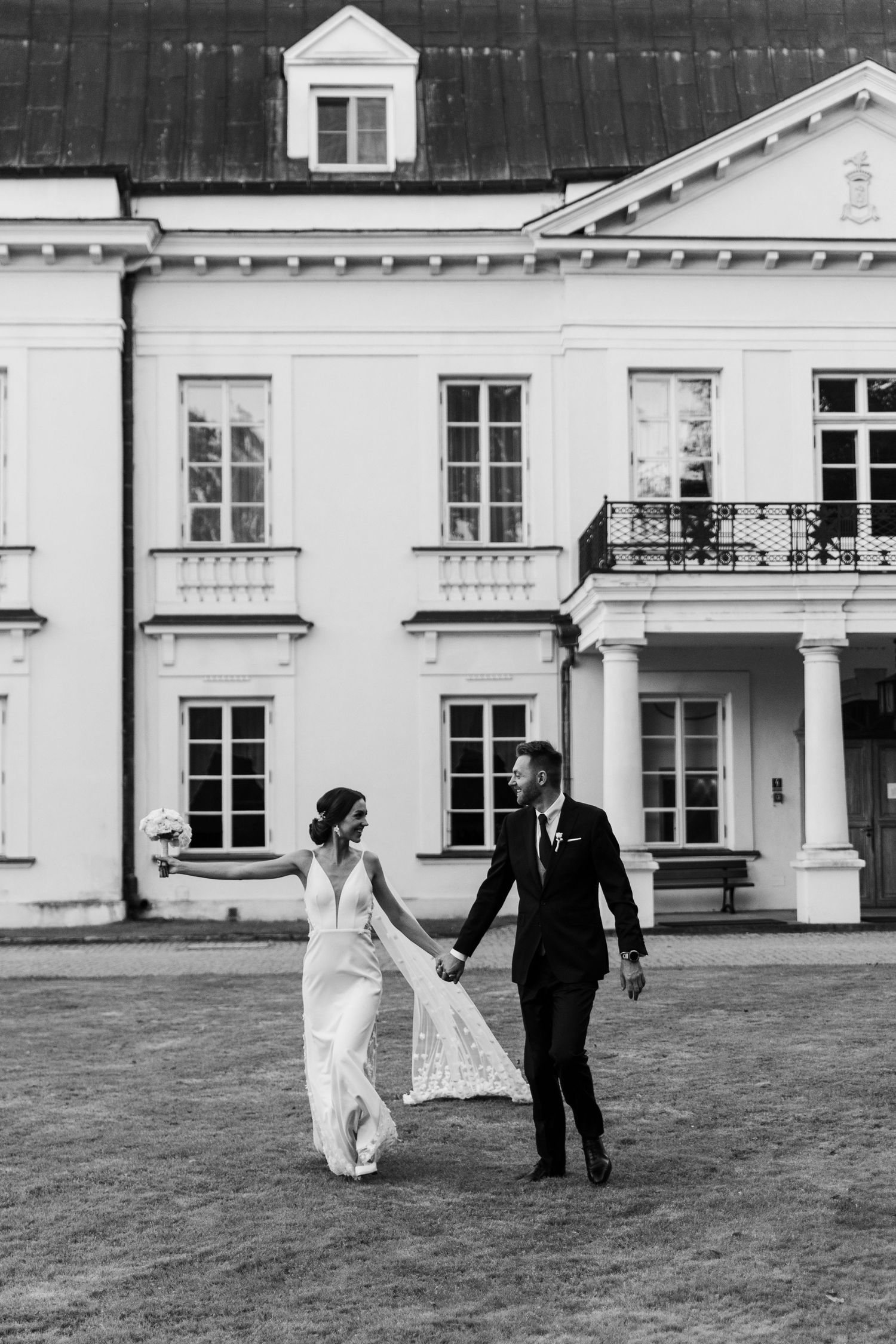 Black and white photo of a couple walking together on a lawn in front of a grand white historic mansion.