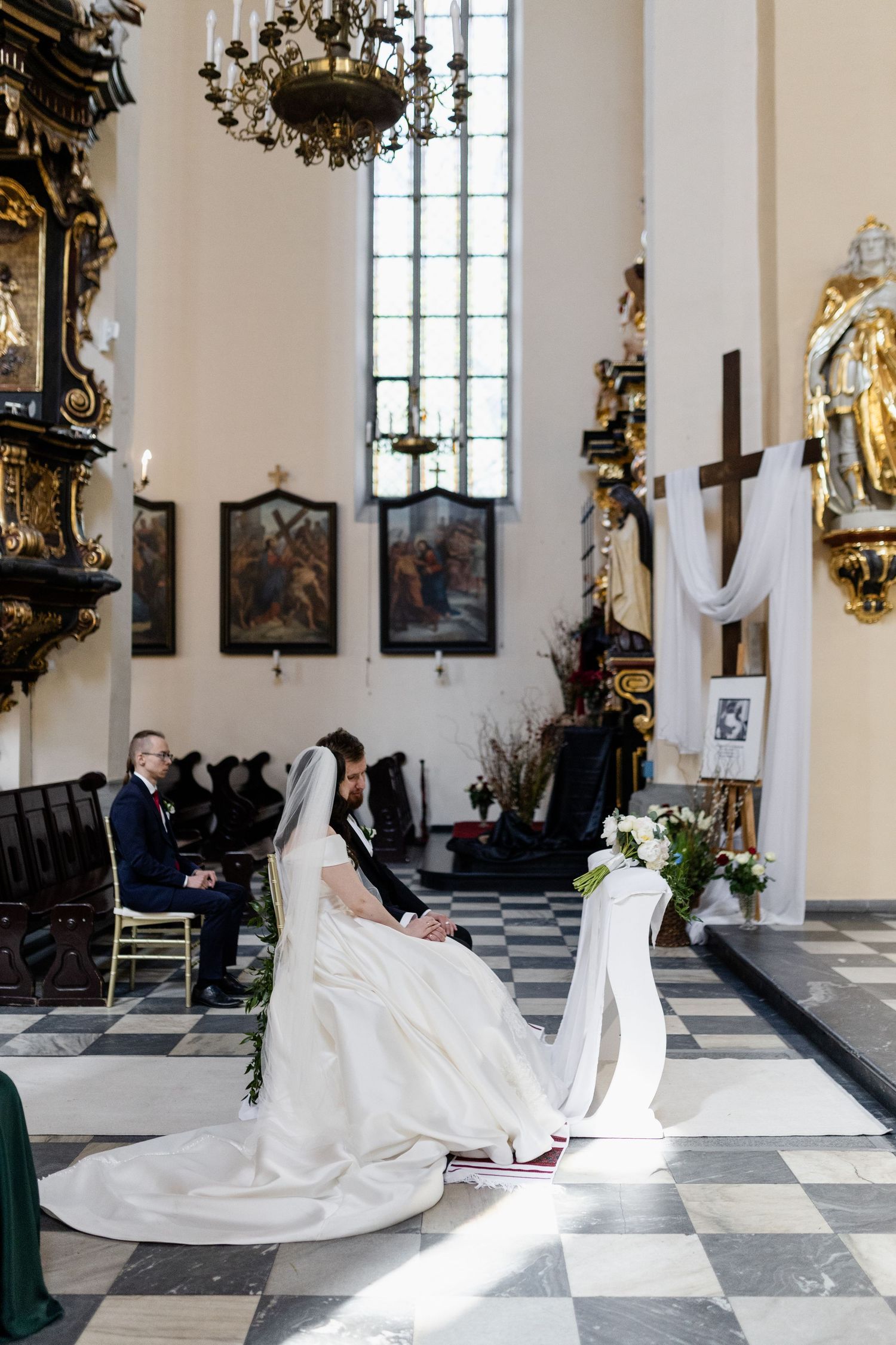 A wedding ceremony inside a historic church with black and white checkered floors and ornate baroque decorations.
