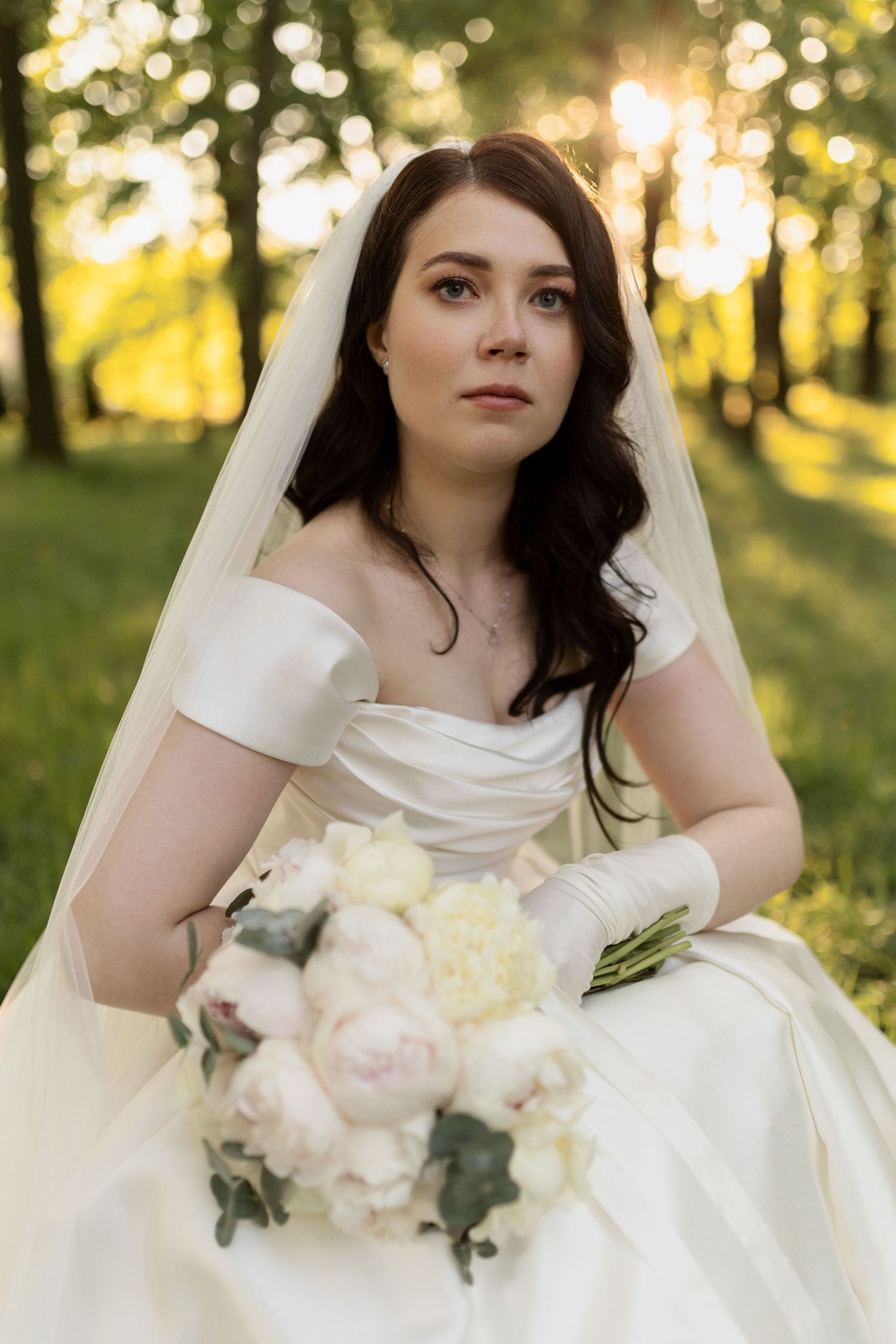 A bride in a white dress with off-shoulder sleeves holds a bouquet of pale flowers while posing in a sunlit forest setting.