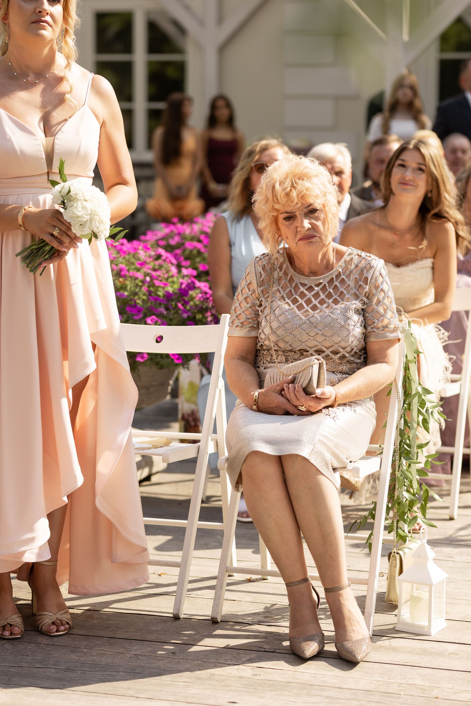 Wedding ceremony guests seated on white chairs with pink flowers in the background at an outdoor venue.