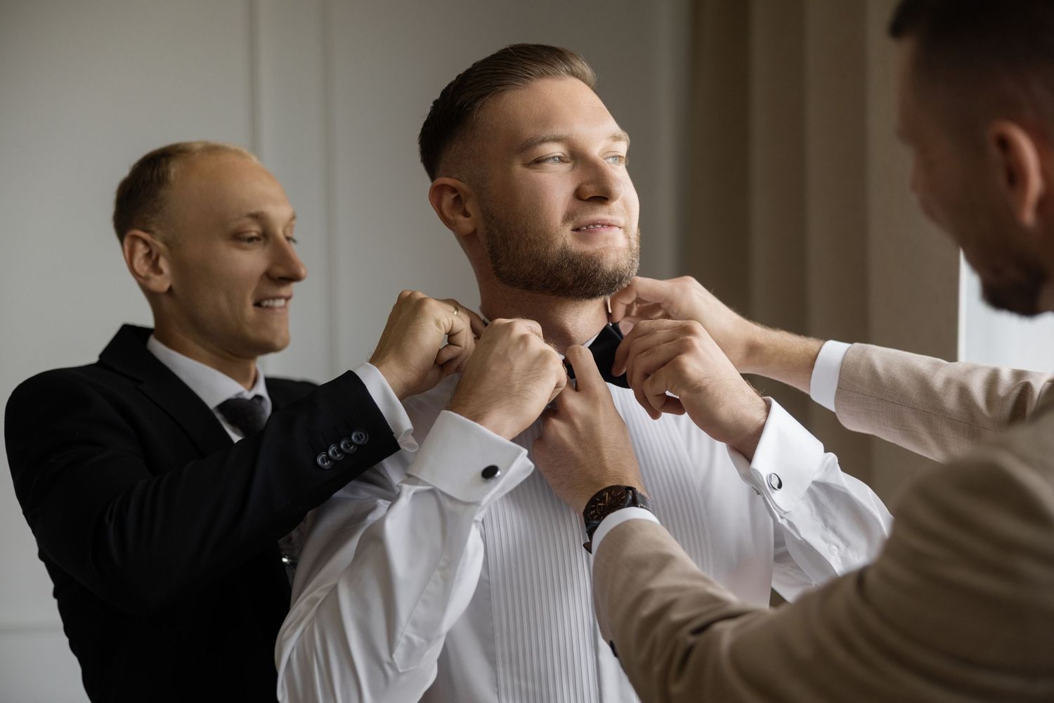 Two groomsmen help a groom adjust his bow tie while dressing for a wedding ceremony.