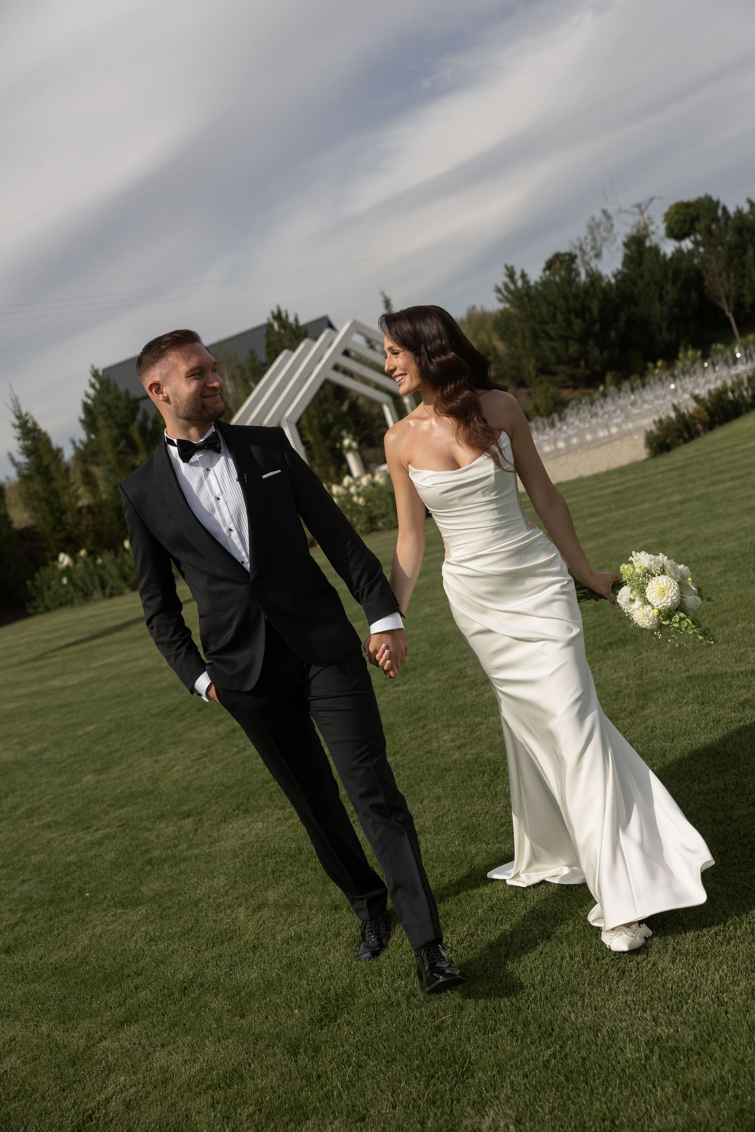 Bride and groom holding hands walking on green lawn, she in white strapless gown, he in black tuxedo with bow tie.