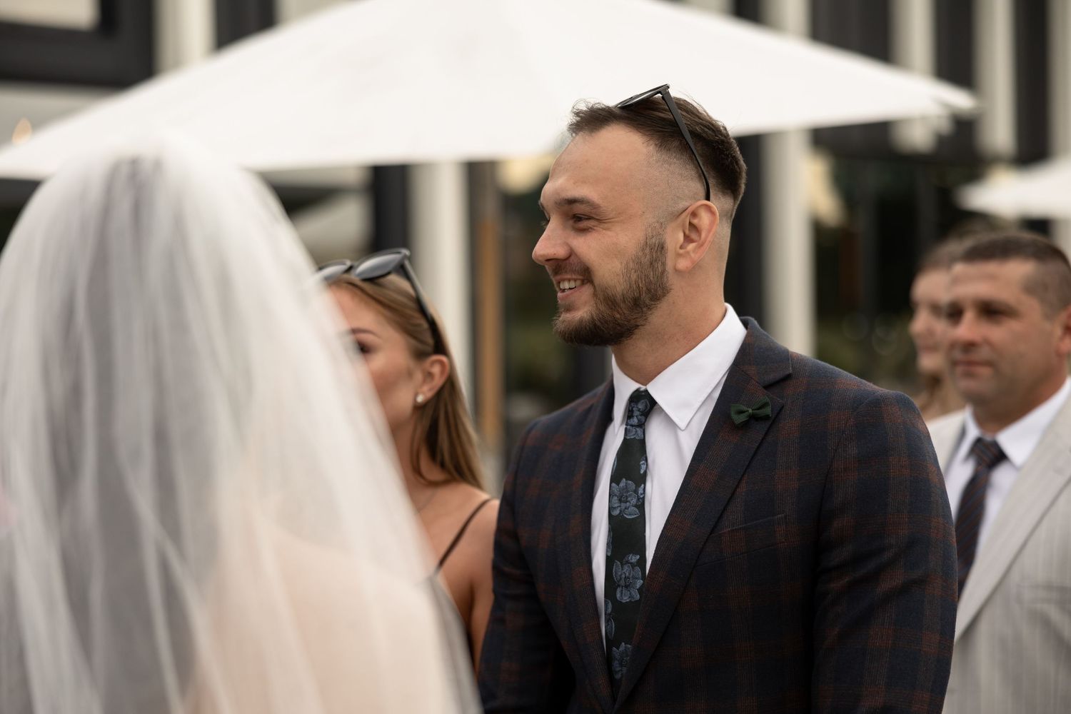 Groom smiling at bride during outdoor wedding ceremony, wearing dark plaid suit with floral boutonniere.