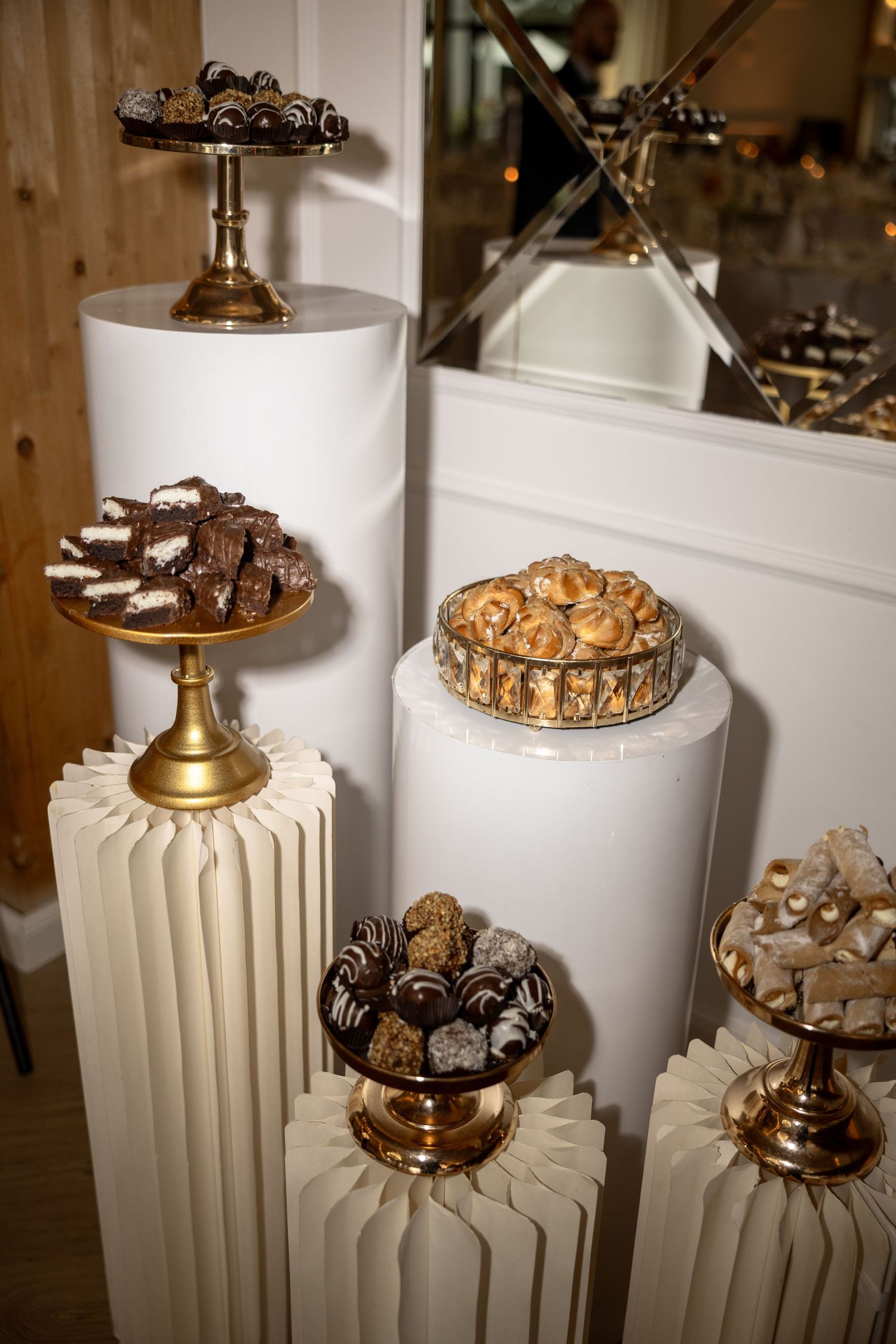 Elegant dessert display with gold stands on white pedestals featuring chocolates, pastries, and truffles at a wedding reception.