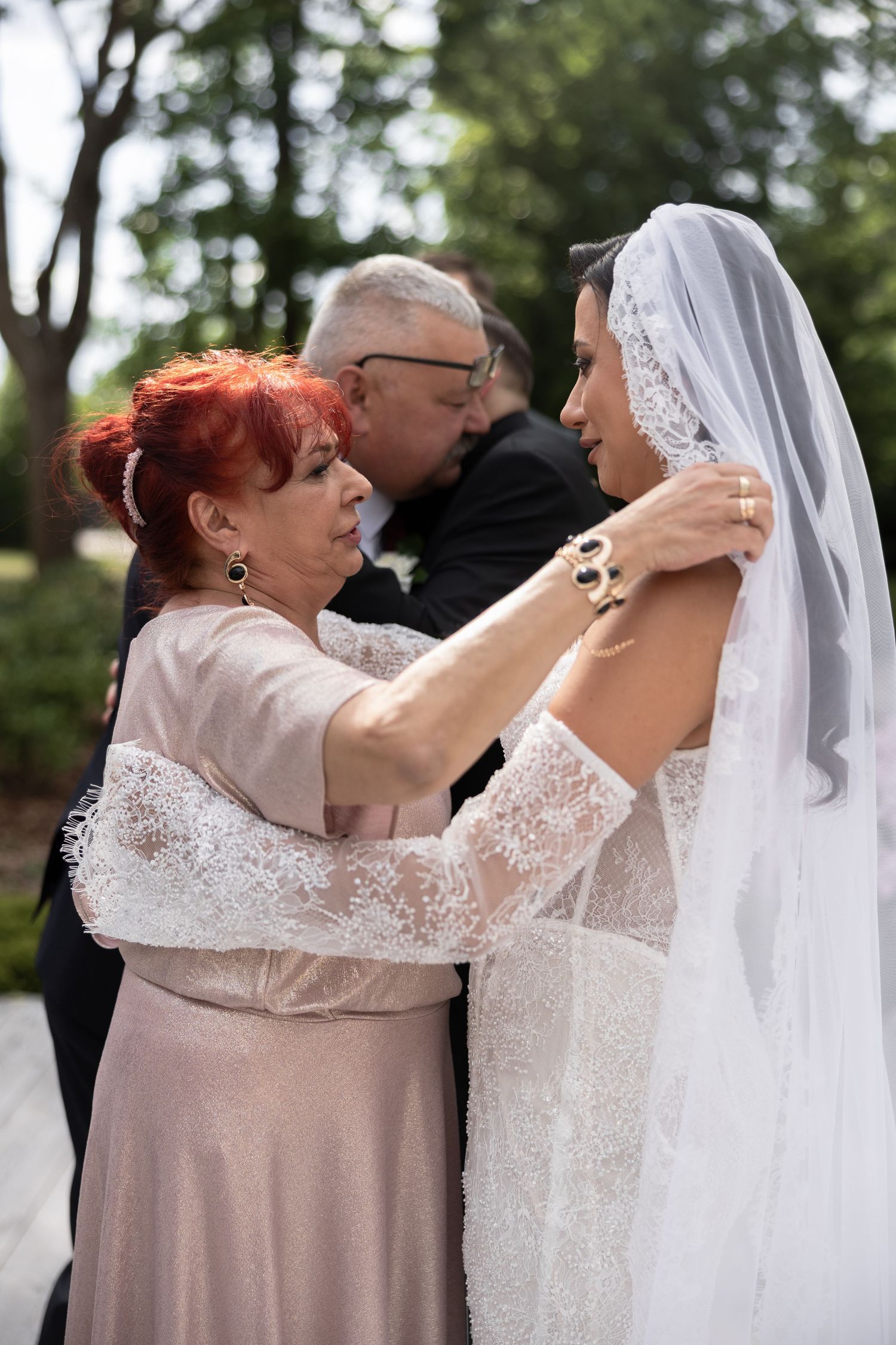 Mother adjusting bride's veil outdoors before wedding ceremony, with older man standing in background.