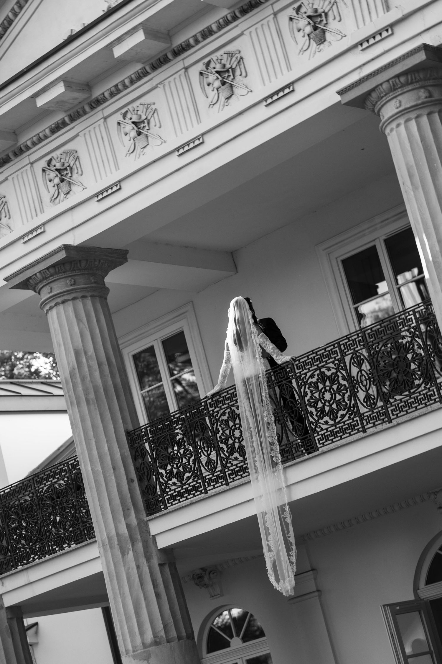 Bride in long veil on ornate wrought iron balcony of elegant white colonial building, black and white photo.