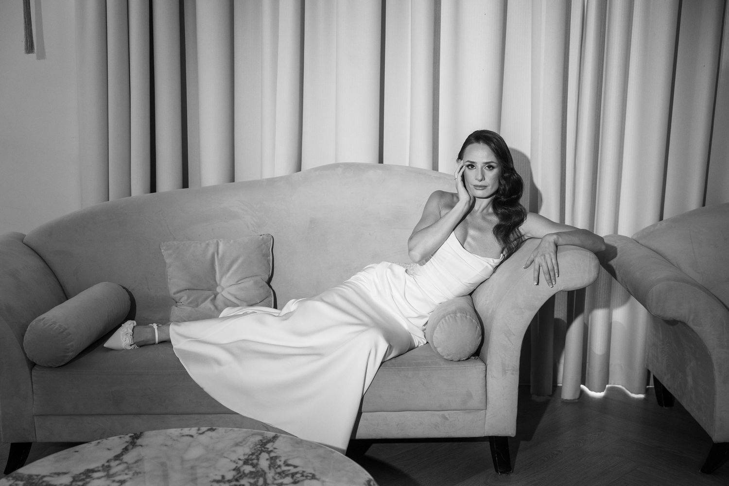 Elegant woman in white gown reclining gracefully on a vintage sofa in a softly lit room, black and white photo.
