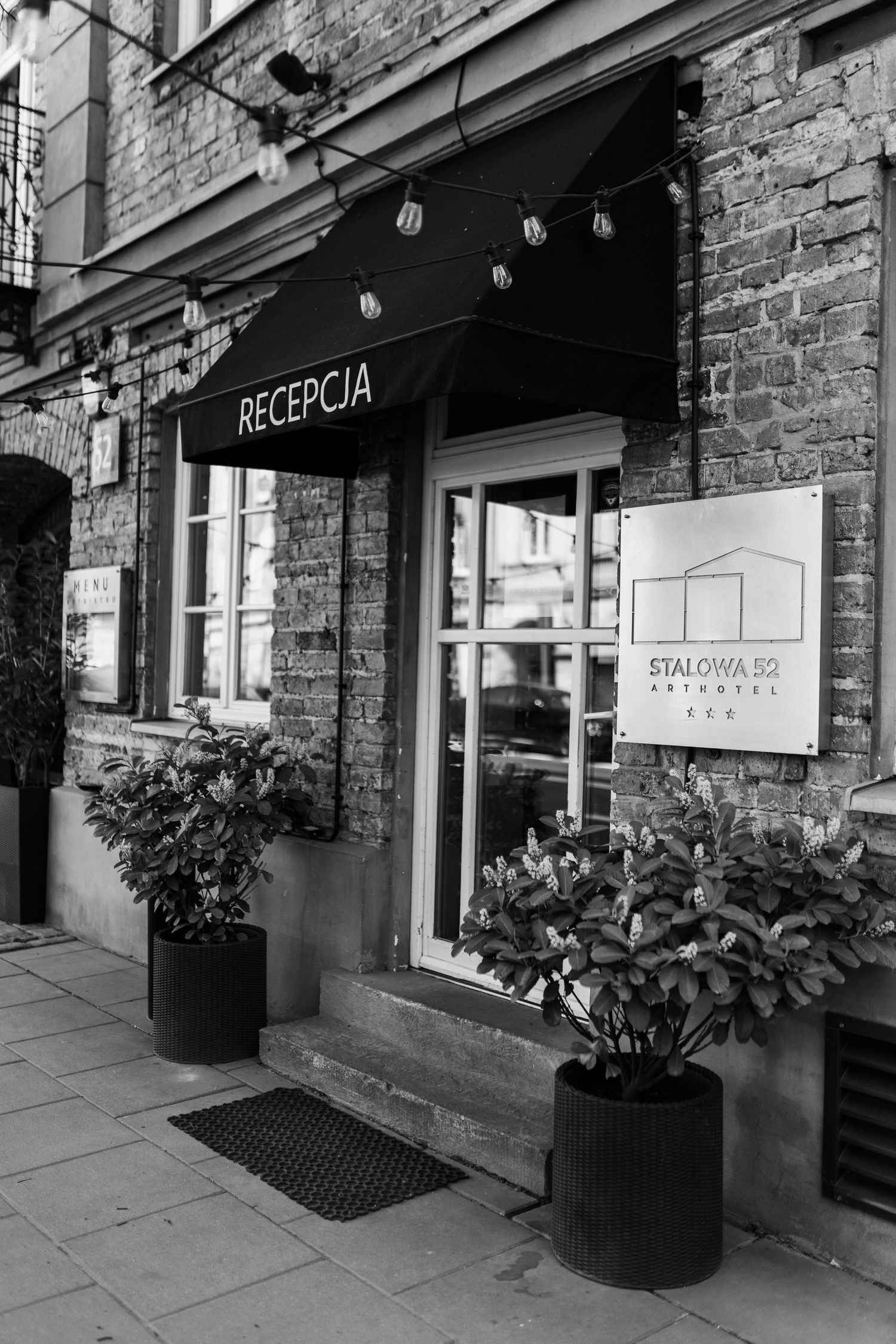 Black and white exterior of a boutique hotel entrance with 'RECEPCJA' sign and decorative potted plants on the sidewalk.