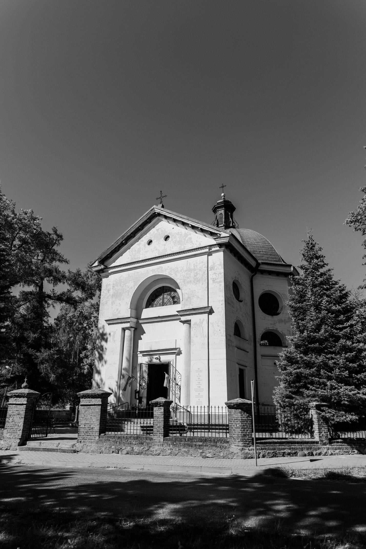 A small white church with an arched entrance and crosses on top stands among evergreen trees.