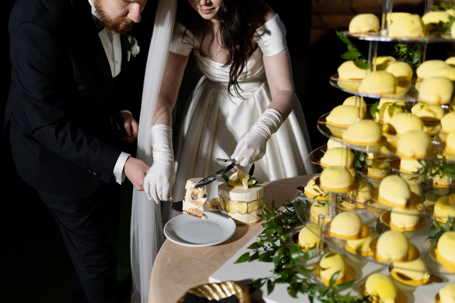 A couple cuts their wedding cake next to a tower of yellow macarons at a formal reception.